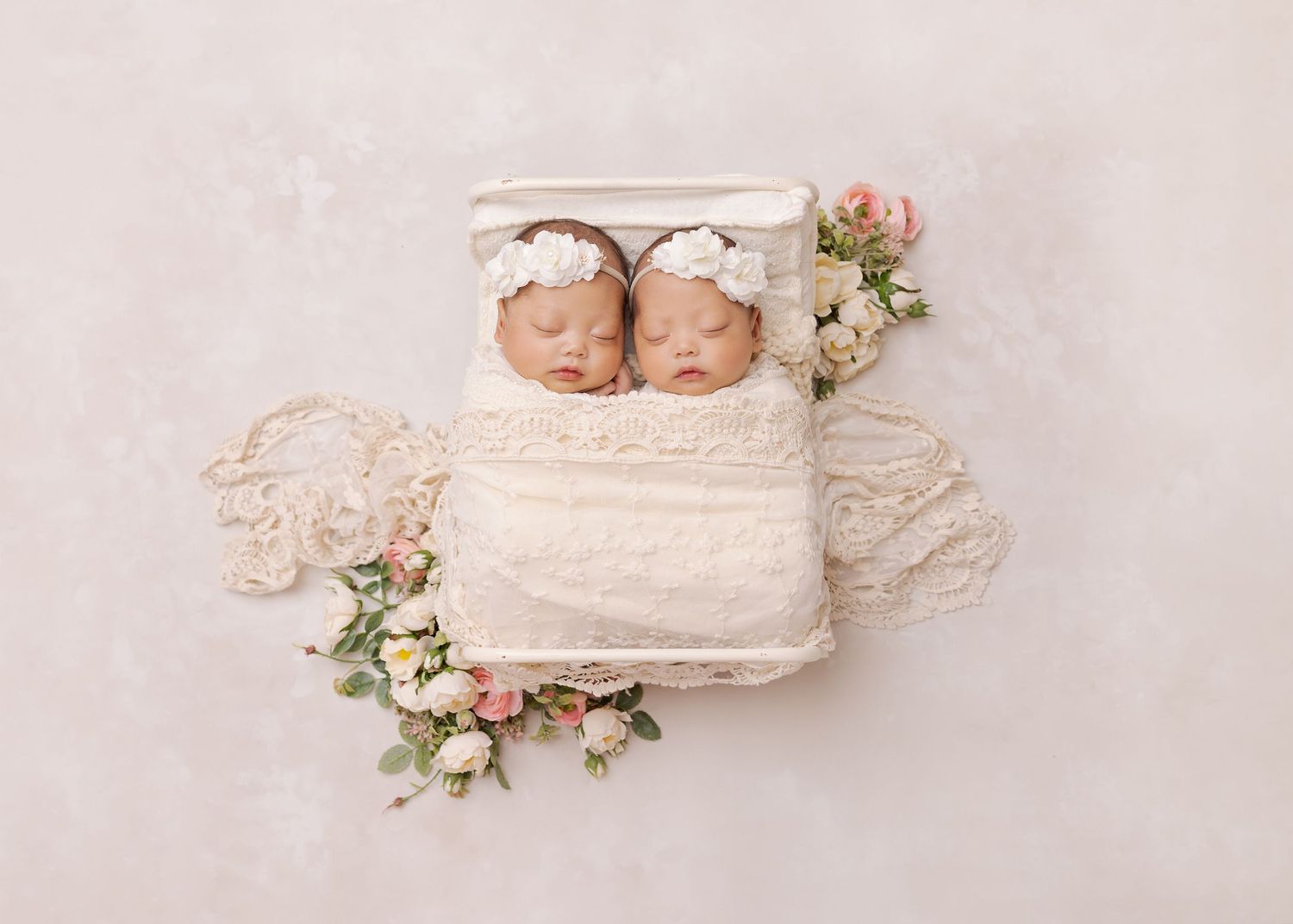 A soft white lace fabric arrangement with baby twins and delicate floral decorations creates a dreamy newborn photo setup.