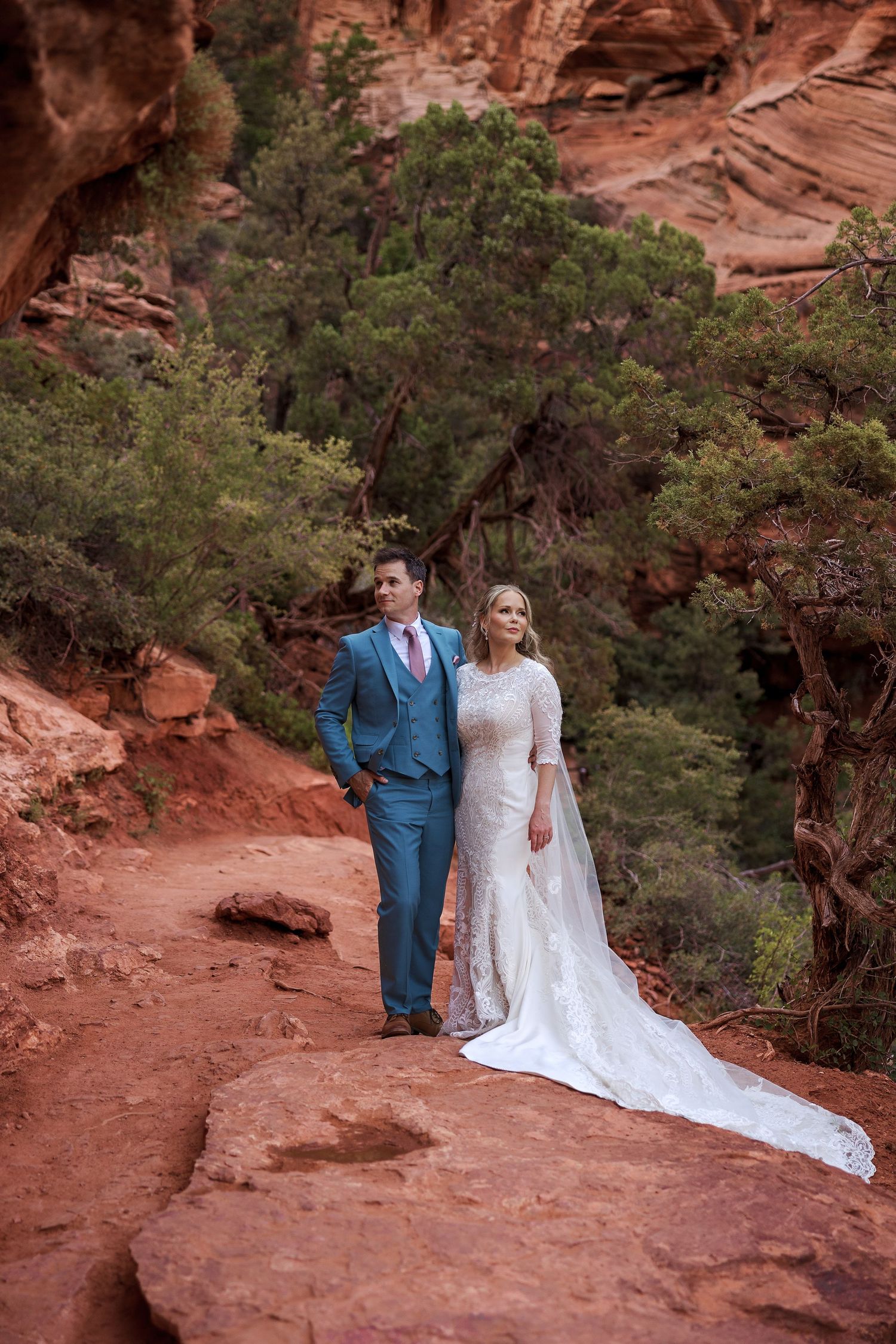 A couple in wedding attire pose on a red rock trail with green trees and canyon walls in the background.