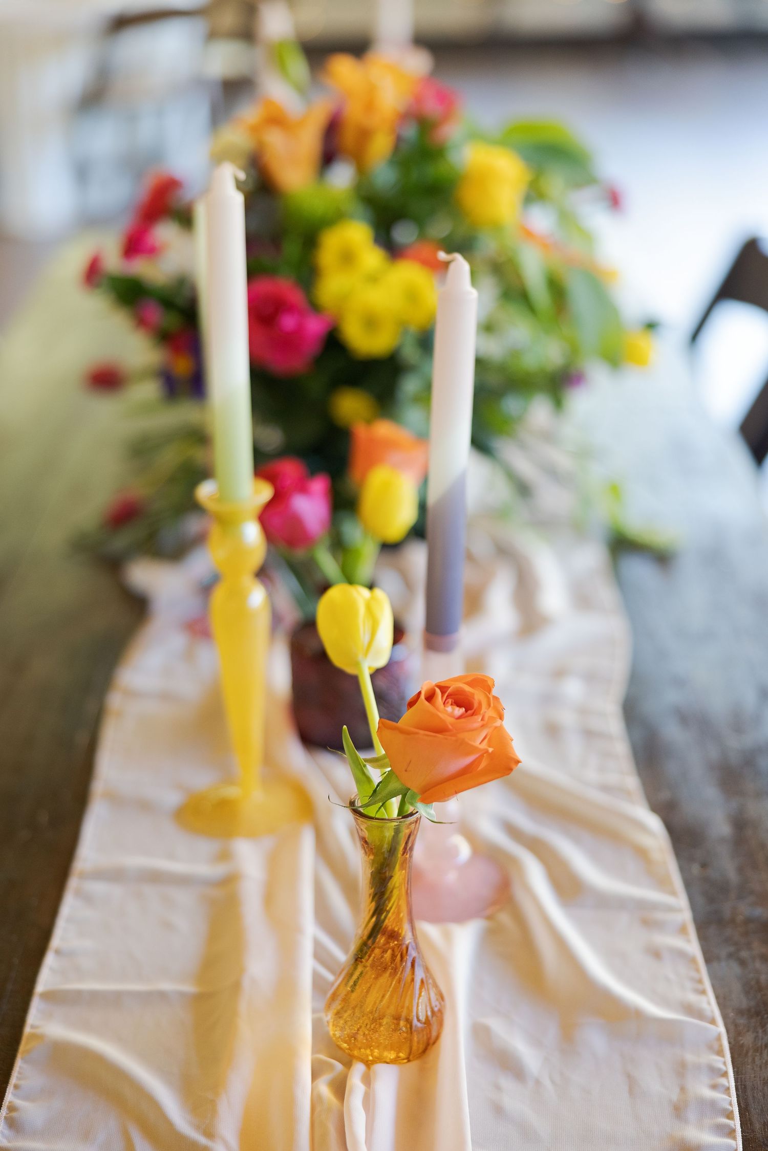 Colorful spring flowers in delicate glass vases with white candles set along a wedding reception table runner.