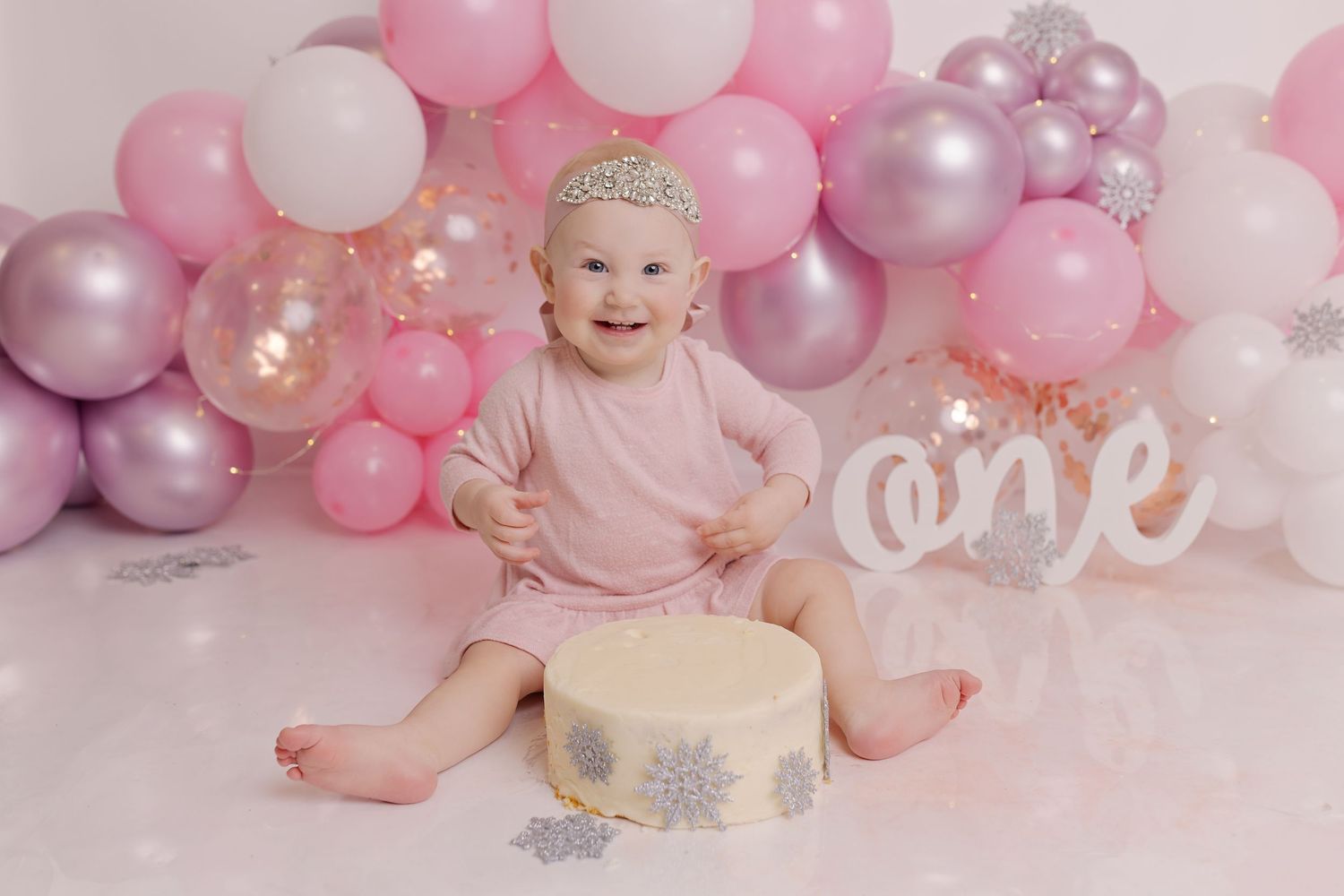 First birthday cake smash photo setup with pink and silver balloons, white 'one' sign, and sparkly cake on pink backdrop.