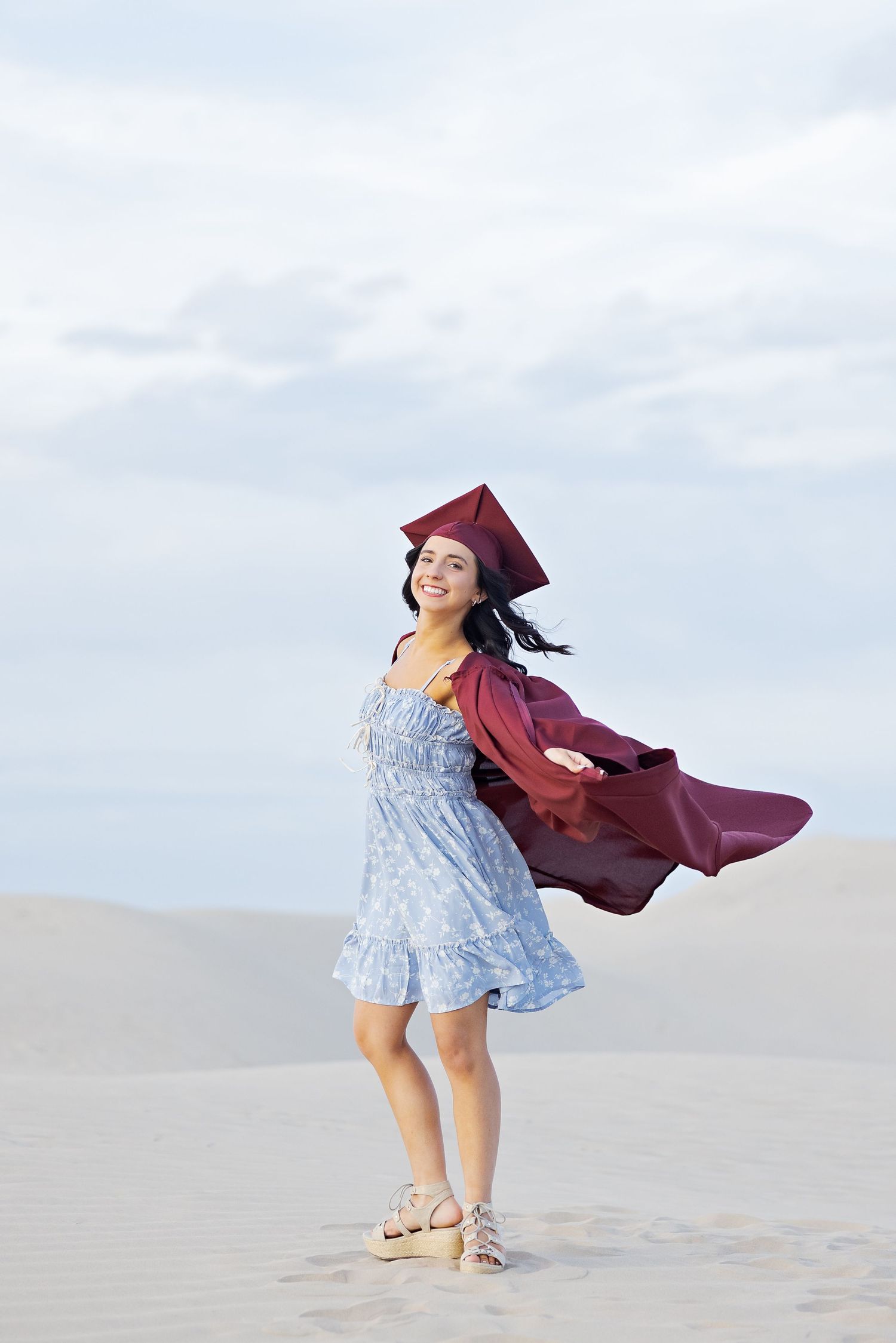 Graduate in burgundy cap and gown twirls joyfully on sandy beach wearing light blue dress and sandals.