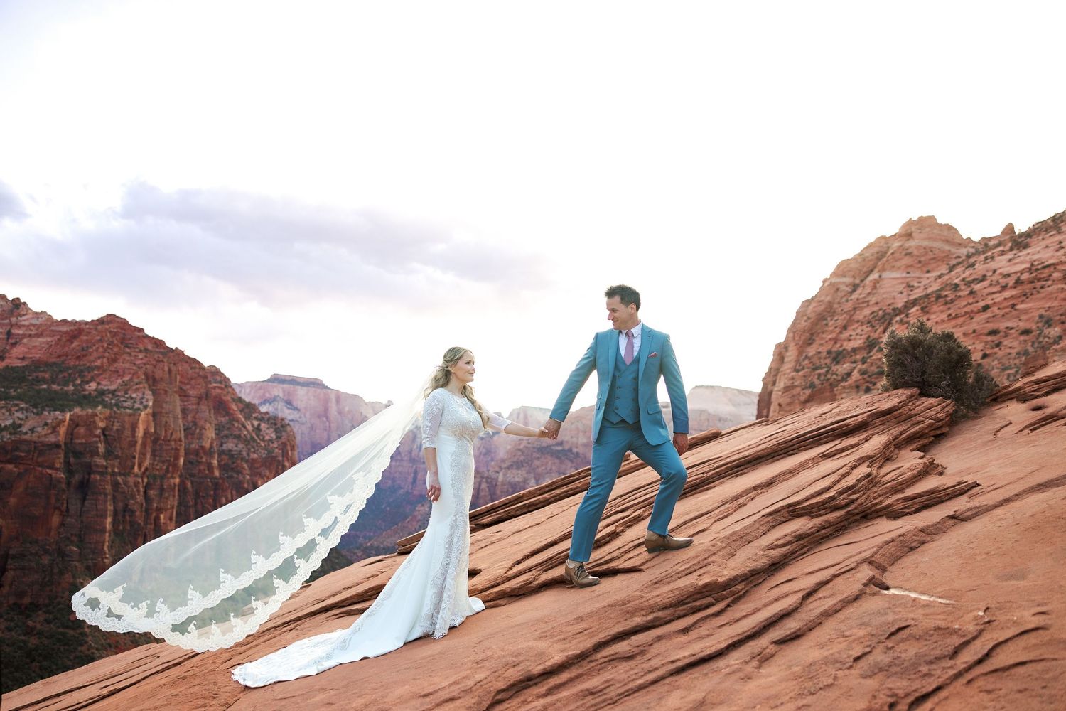 A couple stands atop red rock formations taking photographs at sunset in a scenic desert landscape.