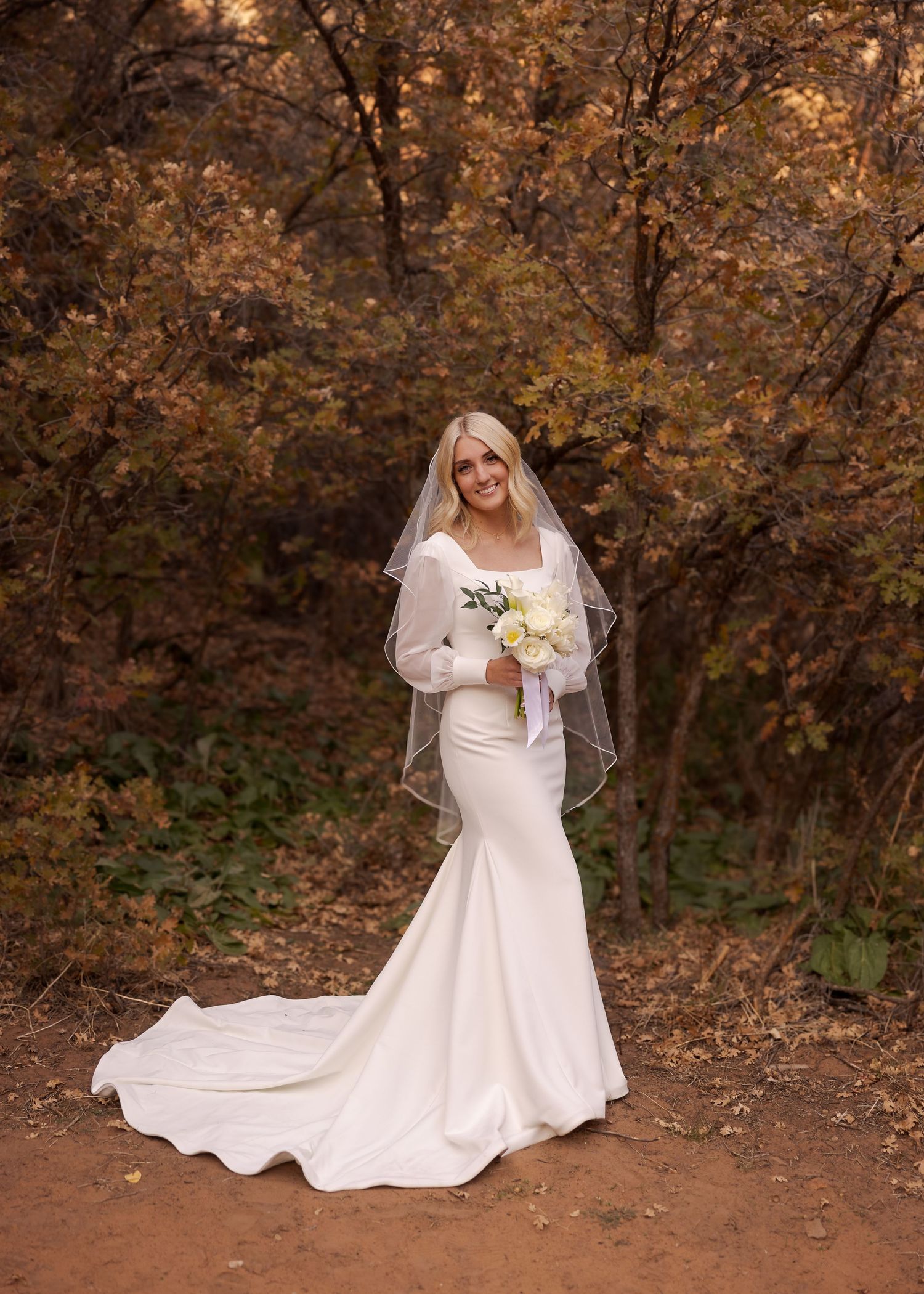 Elegant wedding dress with flowing train and bouquet photographed in autumn woodland setting with golden leaves.