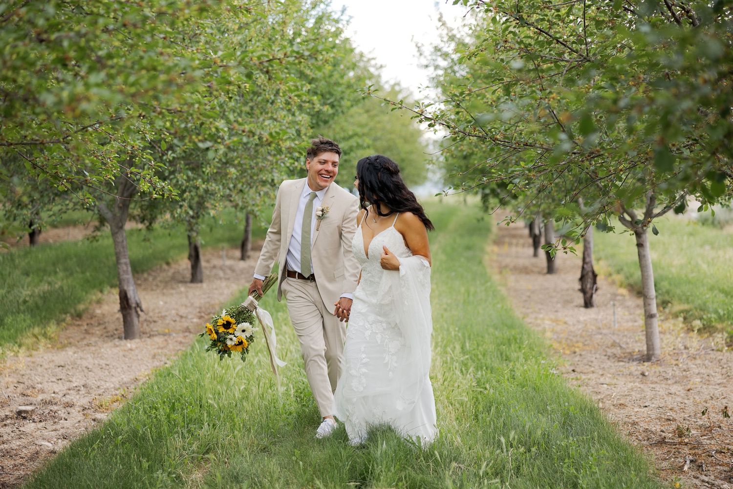 Wedding couple walking through orchard in light colored attire.