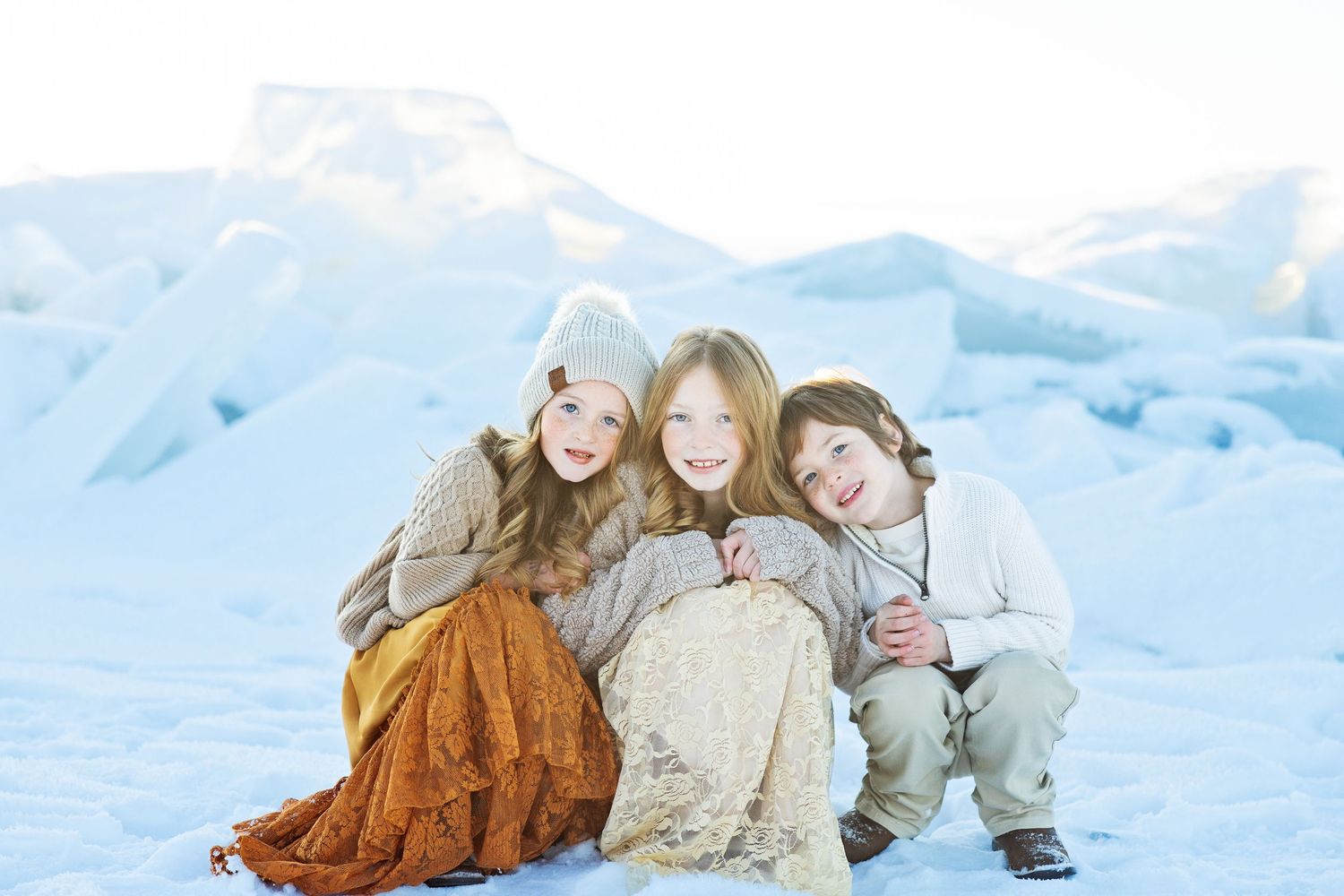 Three people in colorful dresses sitting close together on snowy ground with mountains behind.