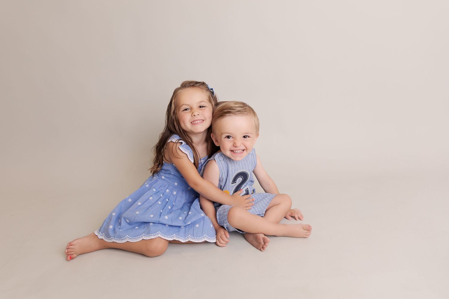 Two young children in blue outfits sitting together on a neutral backdrop.