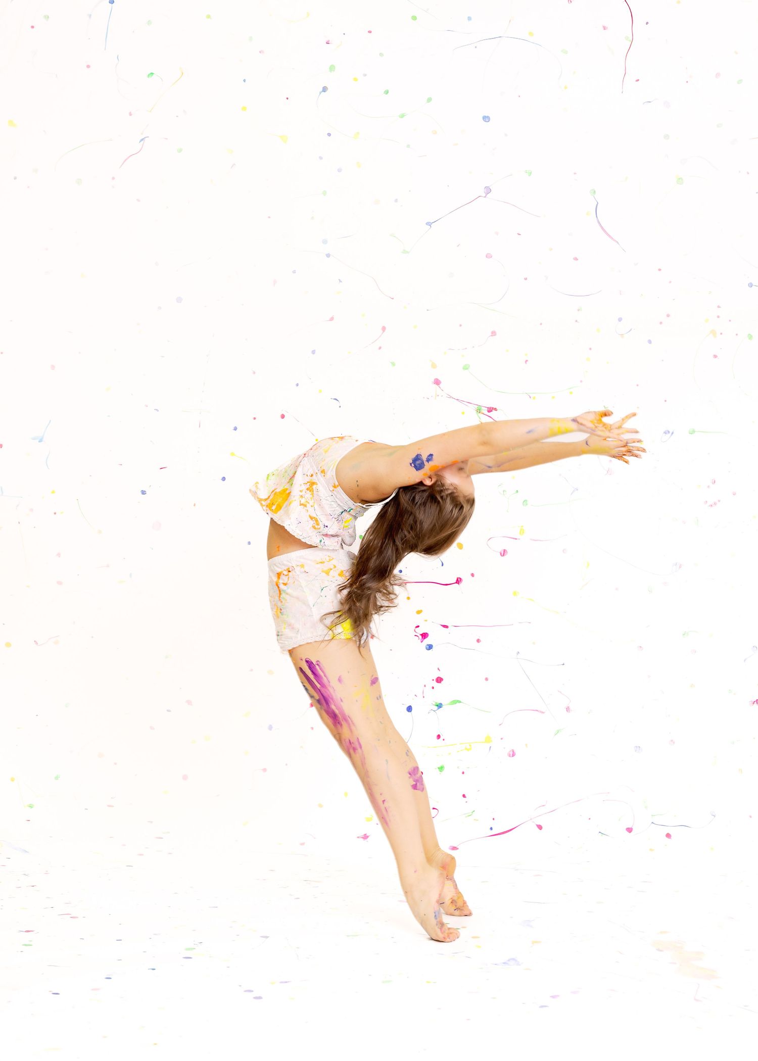Flexible young dancer in paint-splattered outfit performs a backbend leap against a white background.
