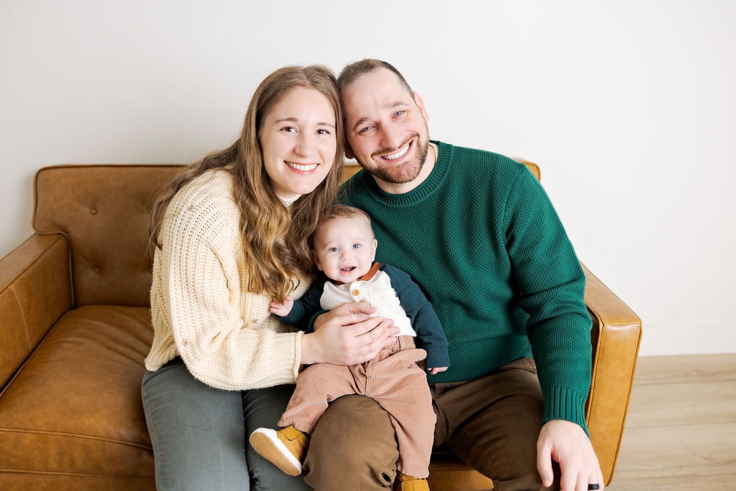 Happy young family with mother, father, and baby sitting together on a brown leather couch smiling.
