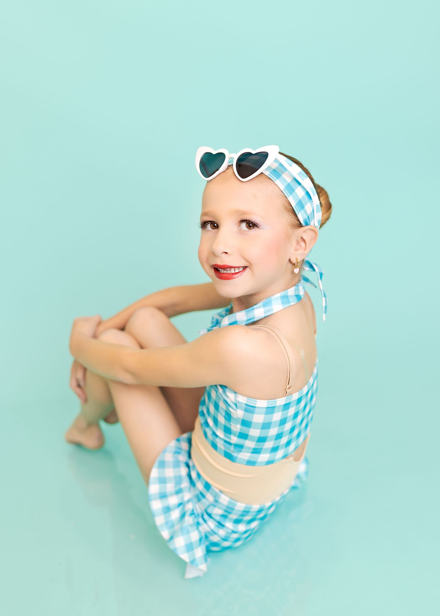 Young girl in teal gingham outfit with heart sunglasses poses on mint background.