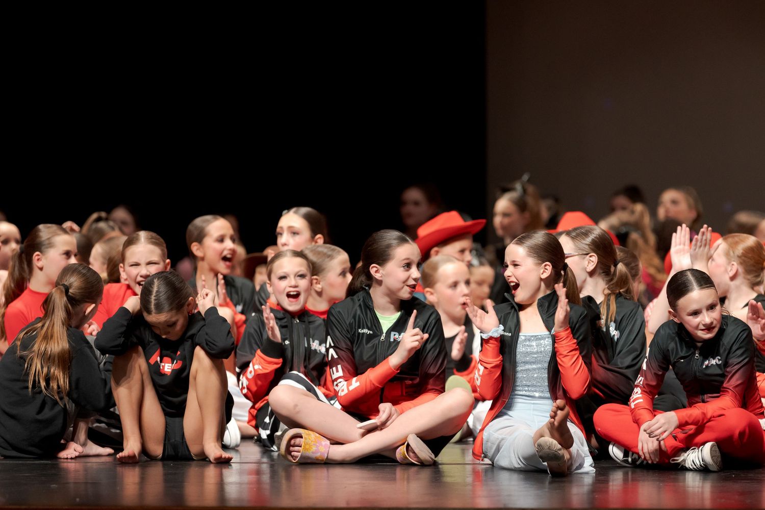 Performers in red and black outfits sit together on stage during a dance recital performance.