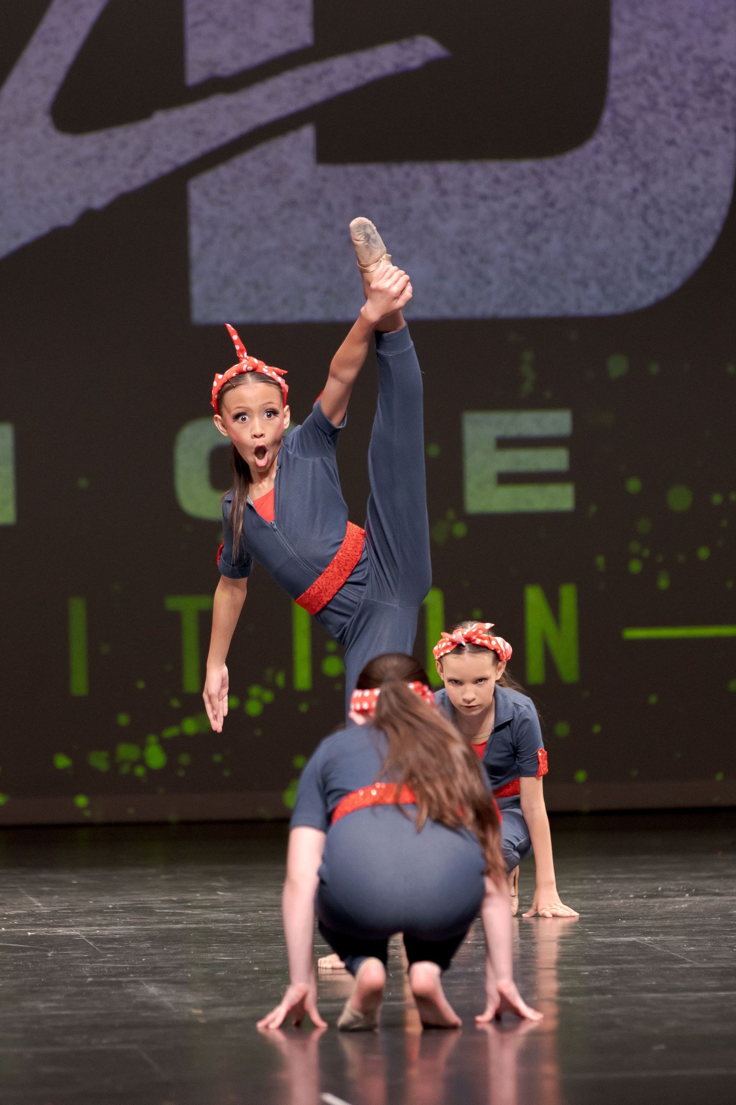 Dancers in navy blue costumes perform an acrobatic contemporary dance routine on a dark stage with green lighting.