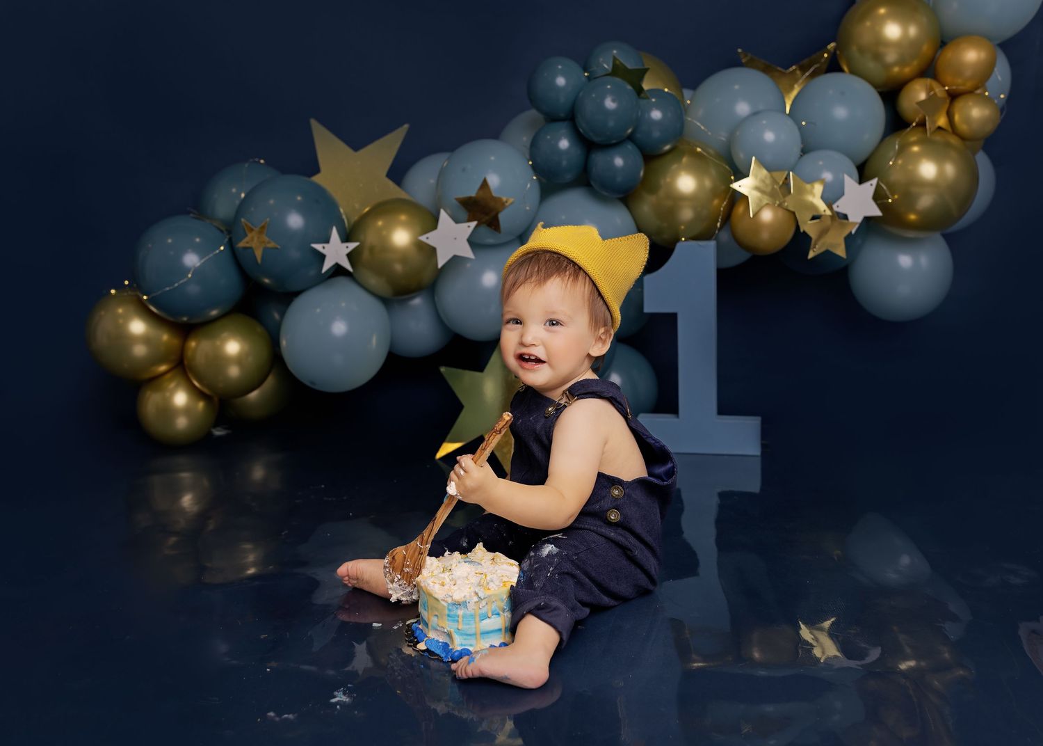 Baby smiling and crawling among gold and blue balloon decorations during a birthday photo session.