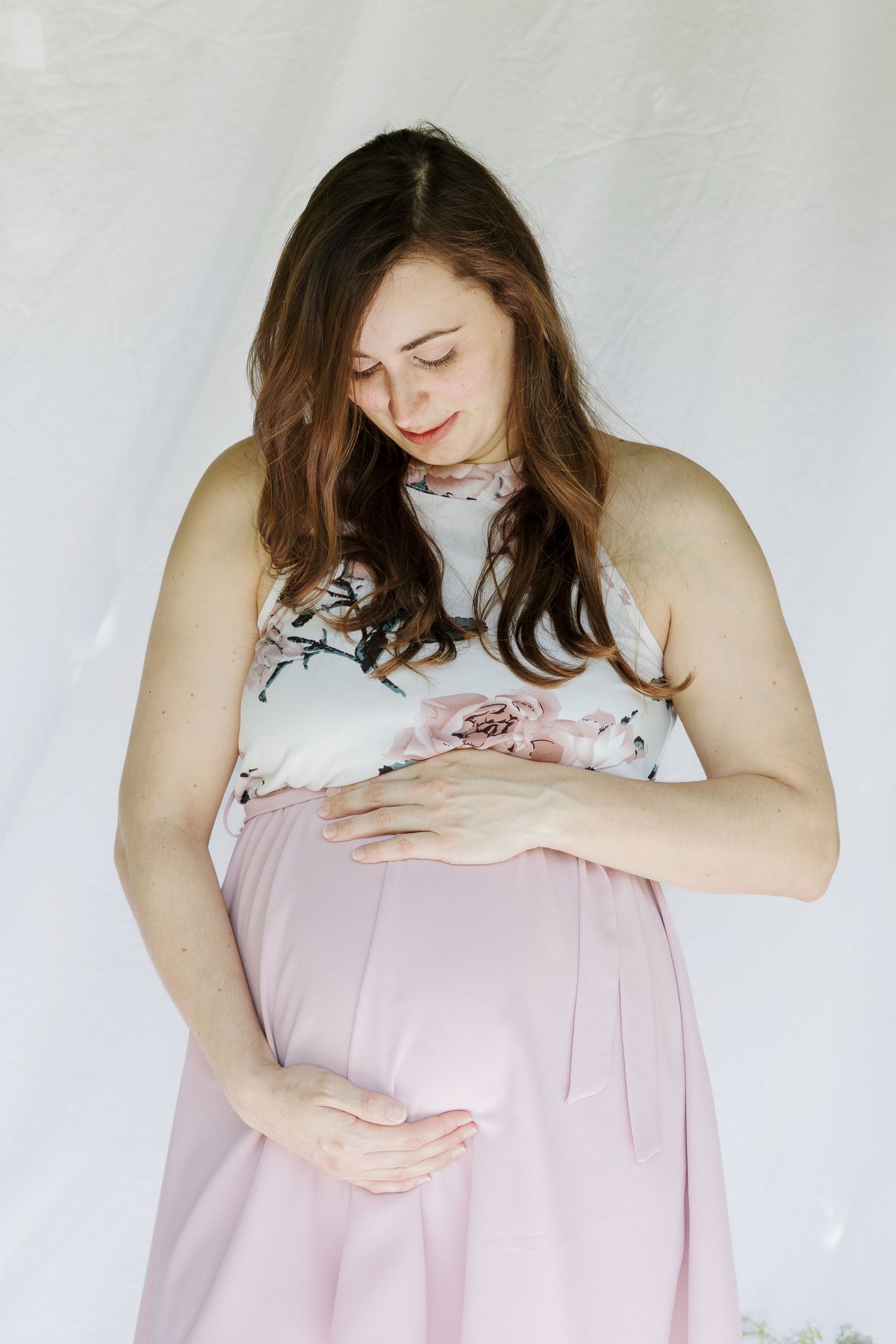 Woman in floral top and pink skirt cradles her pregnant belly against white background.