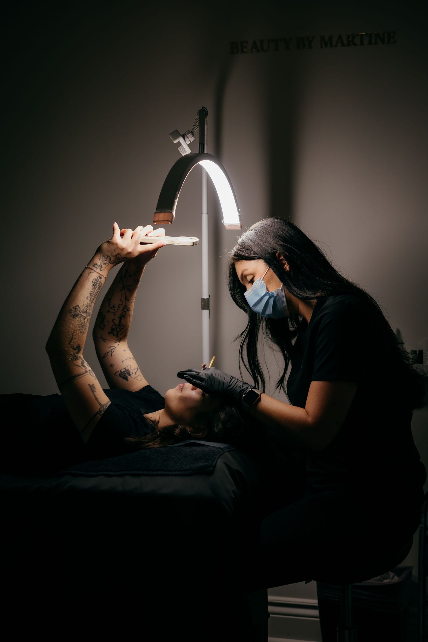 A tattoo artist  works on a client's arm under a bright overhead lamp in a dimly lit studio.