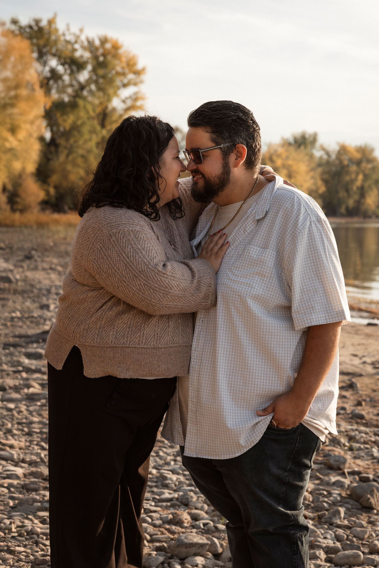 A couple shares an intimate moment on a rocky shore during golden hour as autumn trees glow in the background.