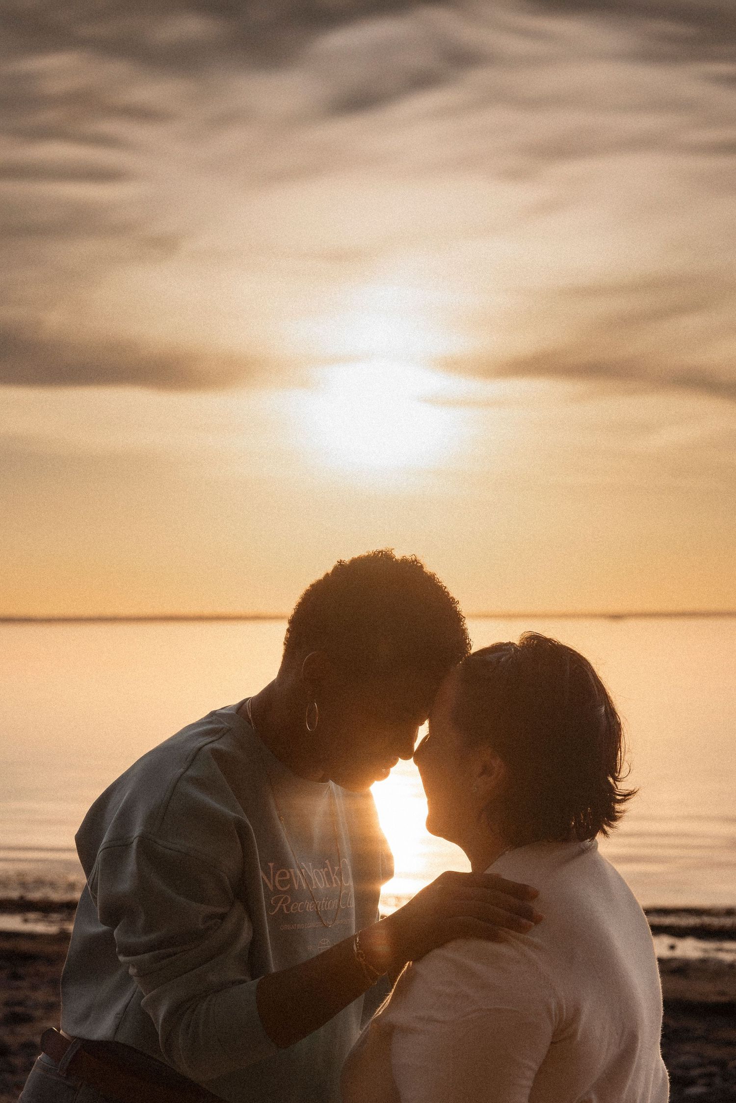 Silhouetted couple shares an intimate moment at sunset by the water's edge in warm golden light.