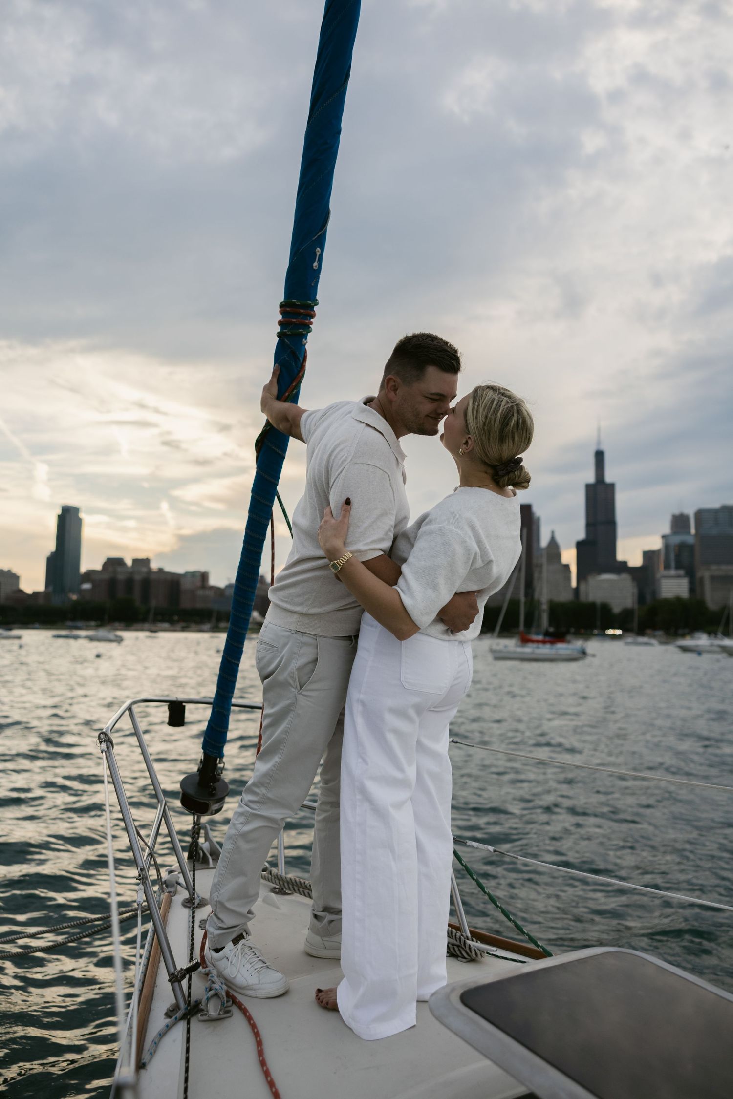 A romantic couple in white outfits embracing on the bow of a sailboat with city skyline silhouetted at dusk.