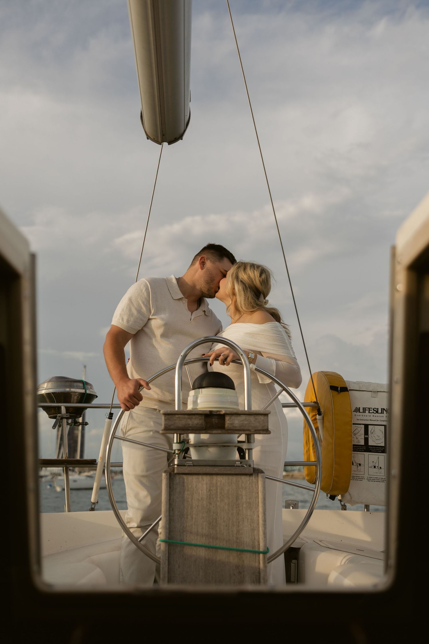A couple in white outfits sharing an intimate moment at the sailboat's steering wheel.