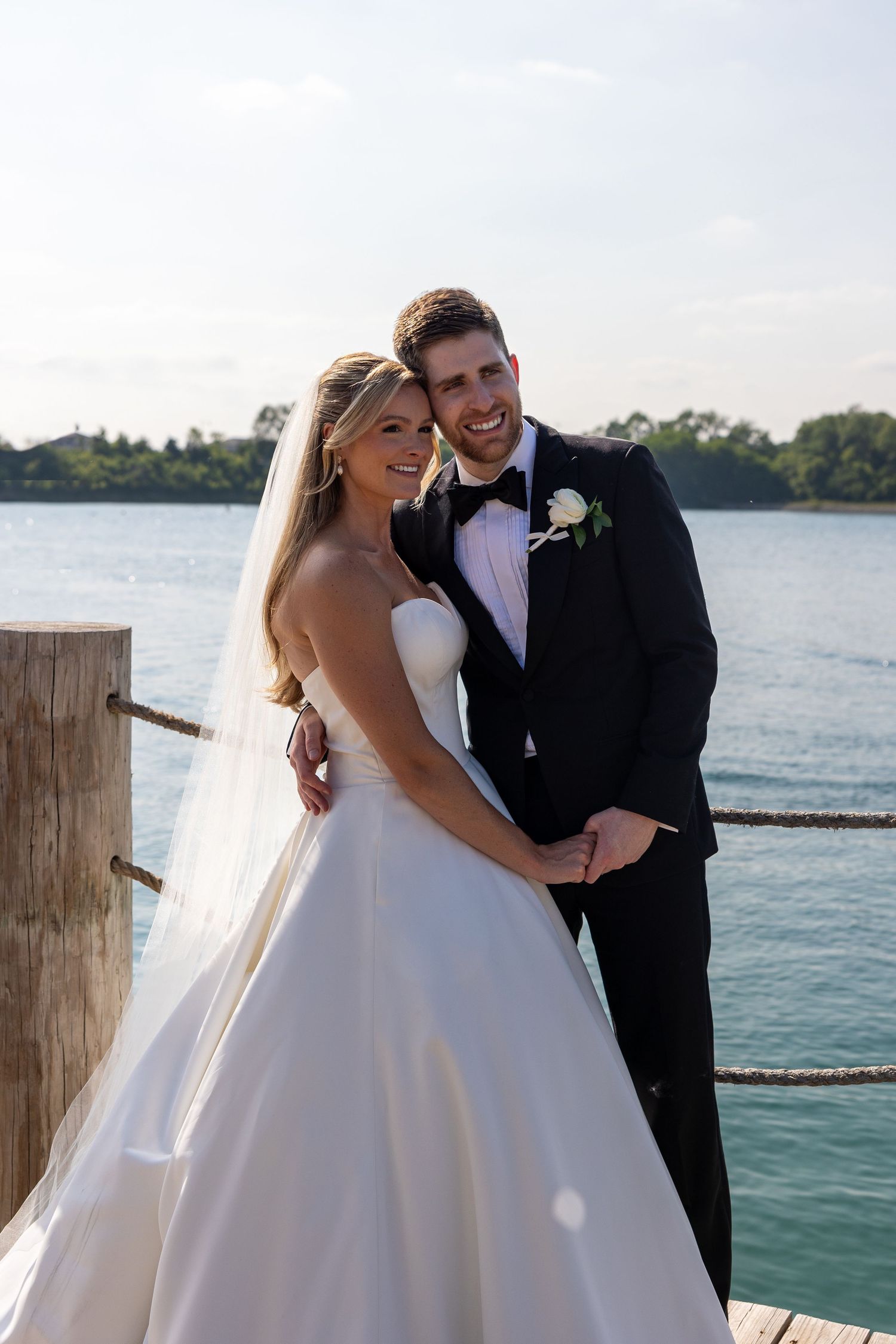 A newlywed couple shares a romantic moment on a dock by a serene waterfront.