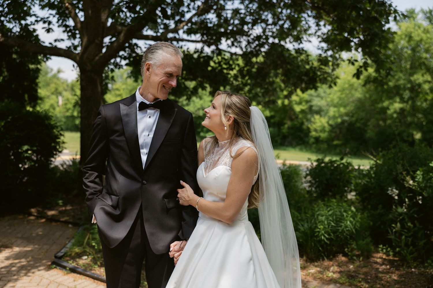 A couple shares an intimate moment outdoors under trees on their wedding day with the bride wearing white.