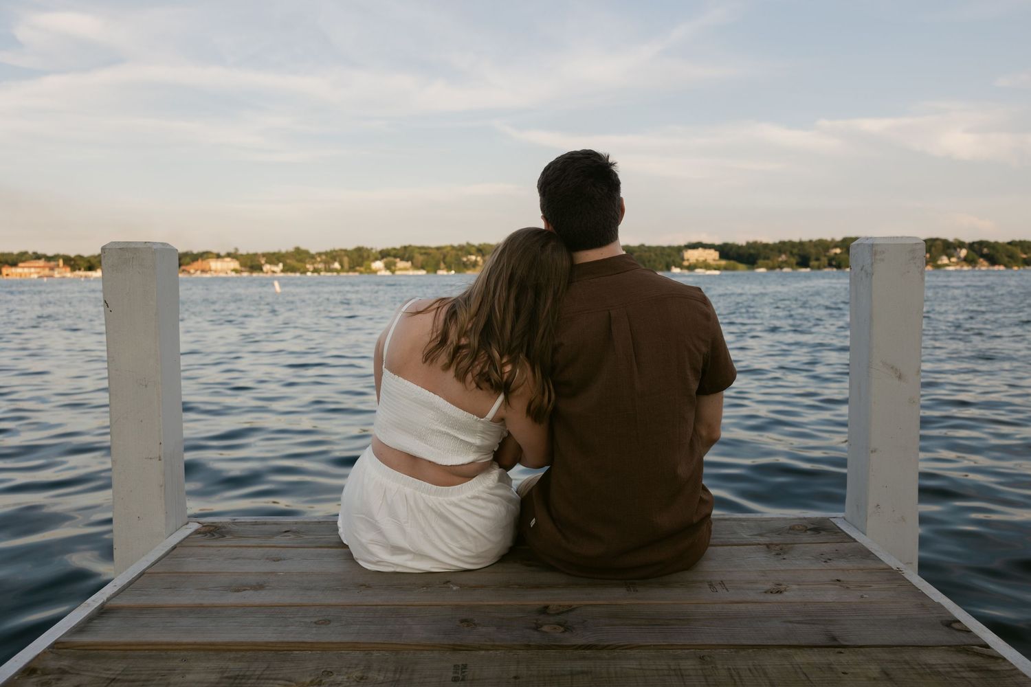 A couple sits close together on a wooden dock overlooking a peaceful waterfront at dusk.