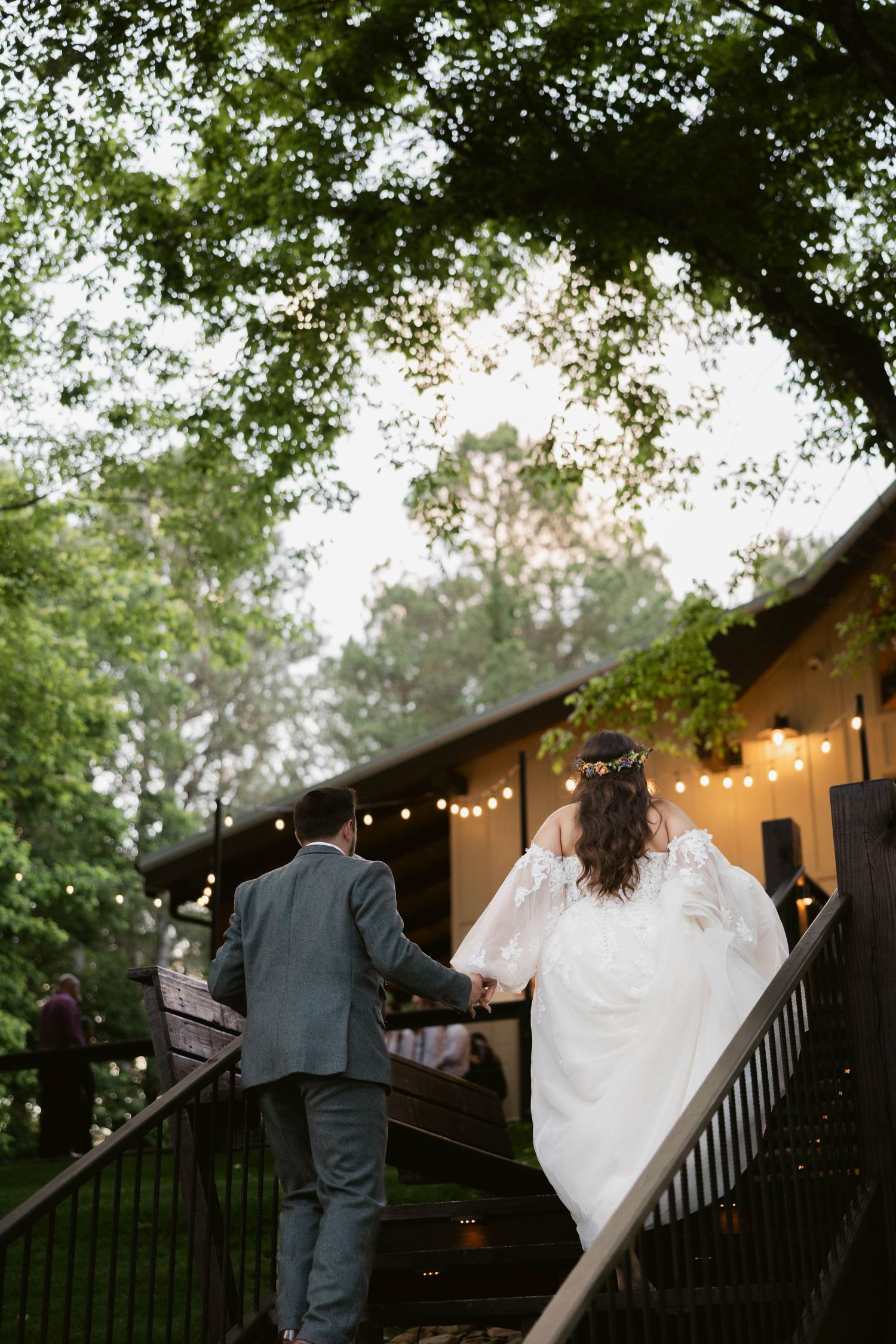 Newlyweds walk up wooden stairs at outdoor venue with trees and string lights.