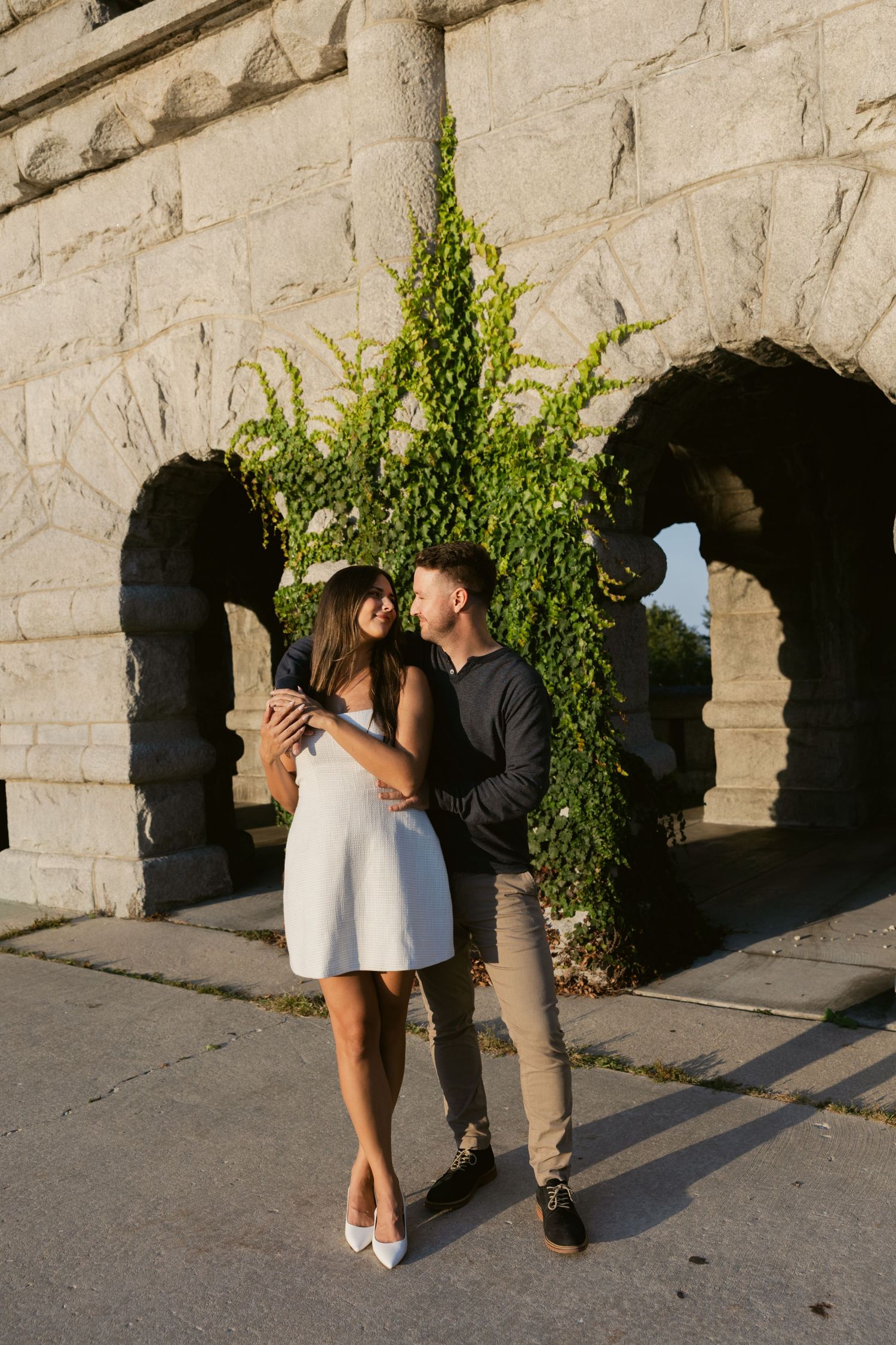 Couple embracing against a stone wall with climbing ivy in warm sunlight.