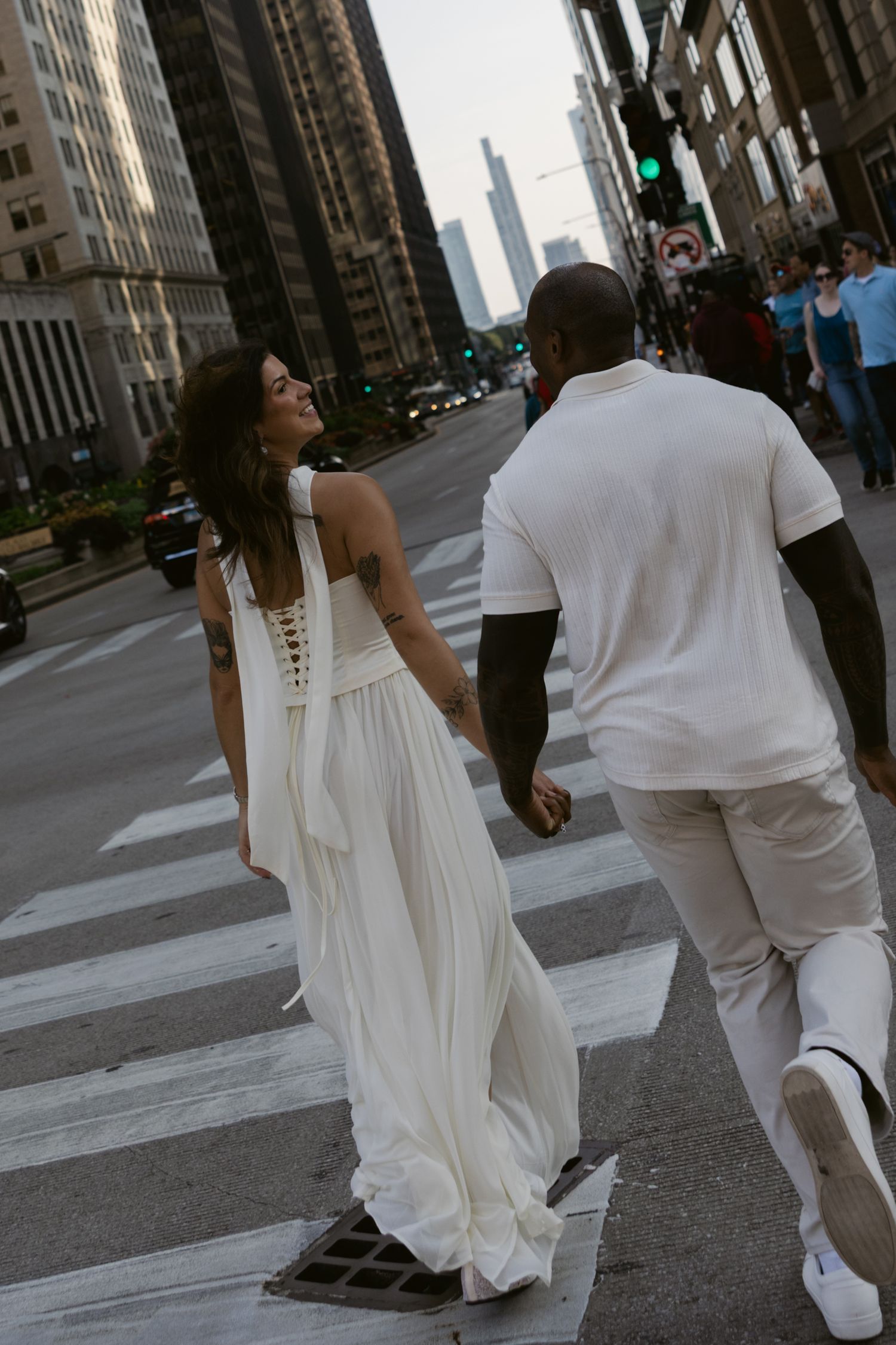 A couple in elegant white attire walks down an urban street with tall buildings in the background.