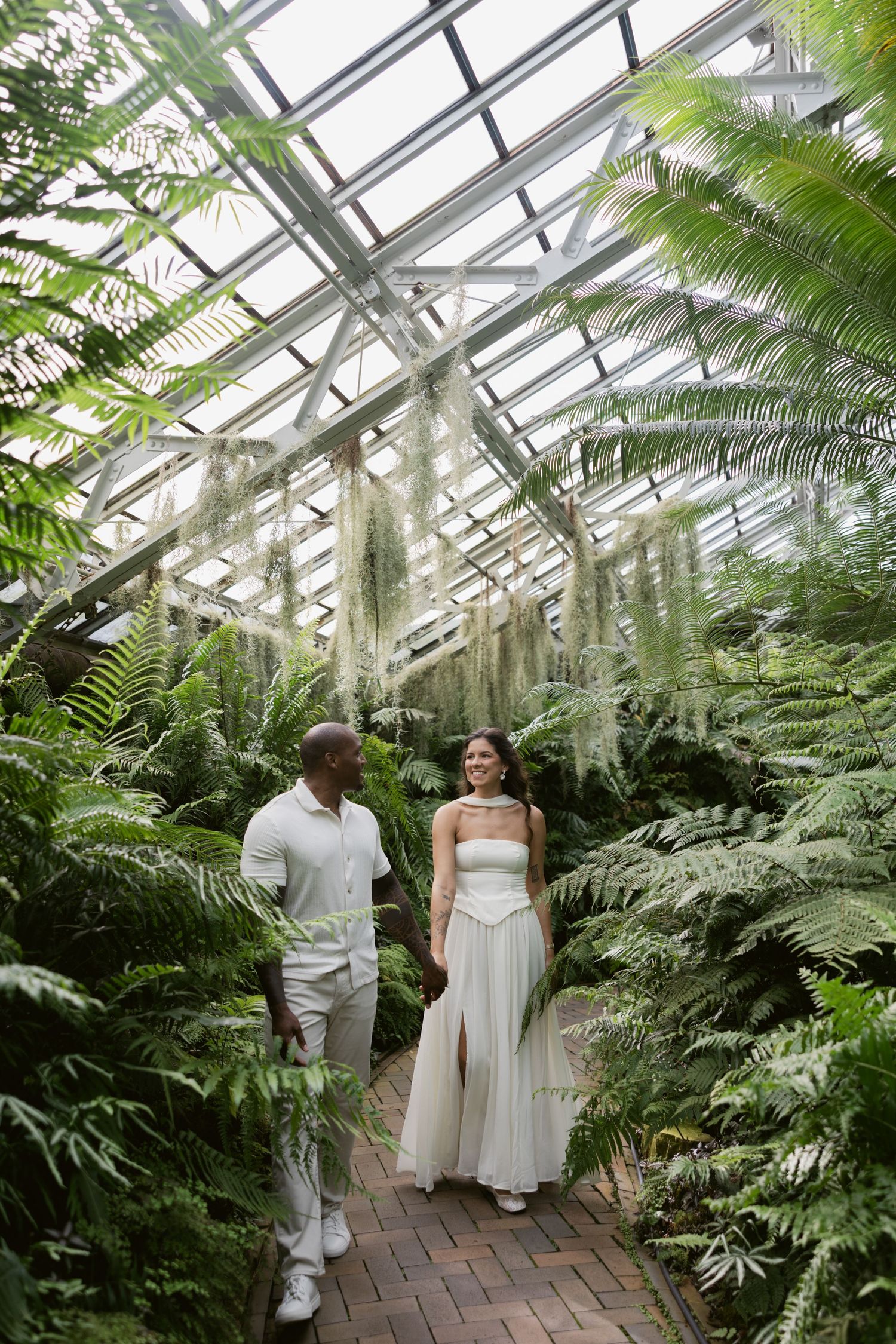 A romantic couple in white attire walks through a lush greenhouse conservatory filled with tropical ferns and plants.