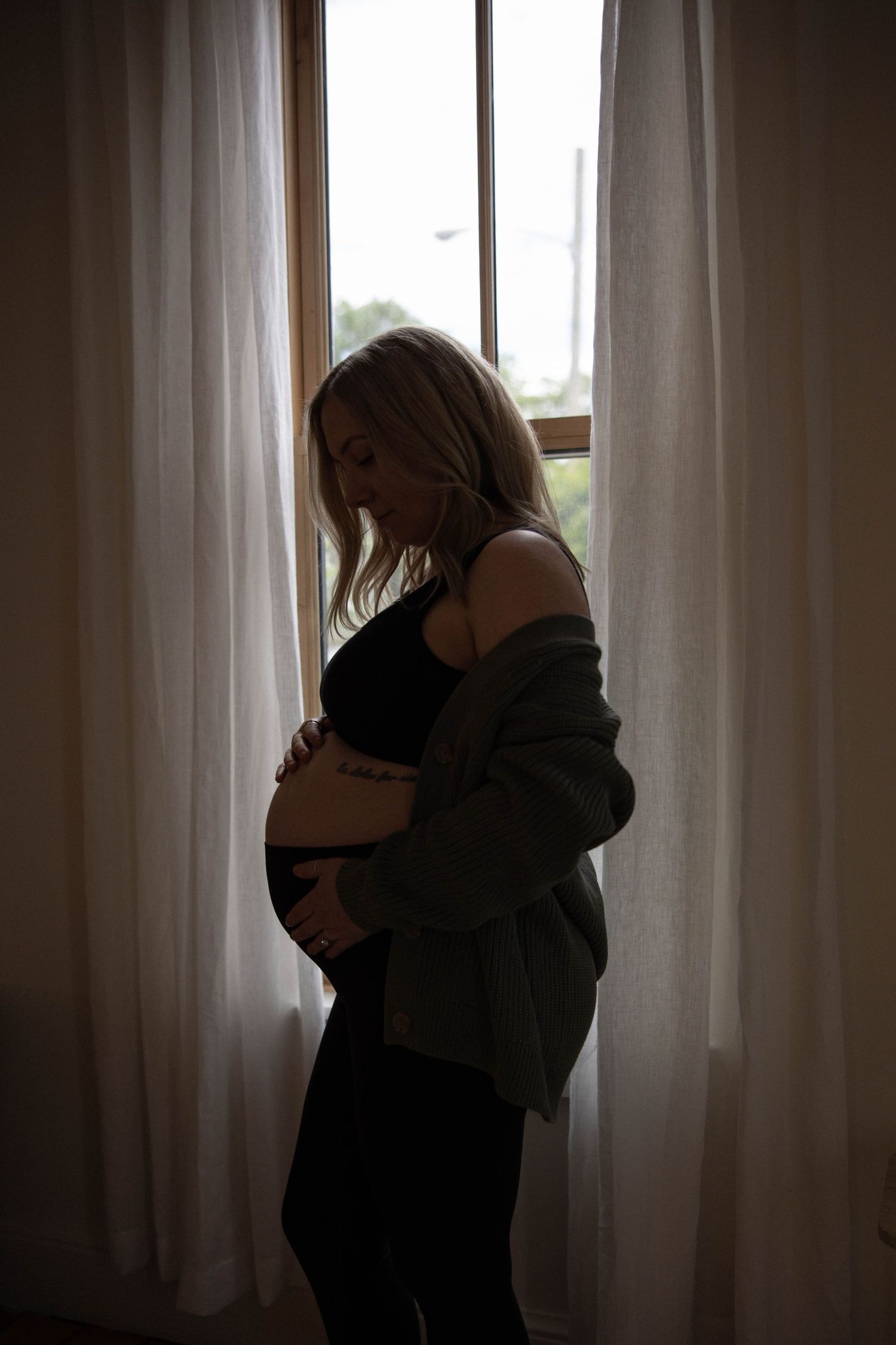 A silhouetted figure stands by a bright window with curtains in an intimate maternity moment.