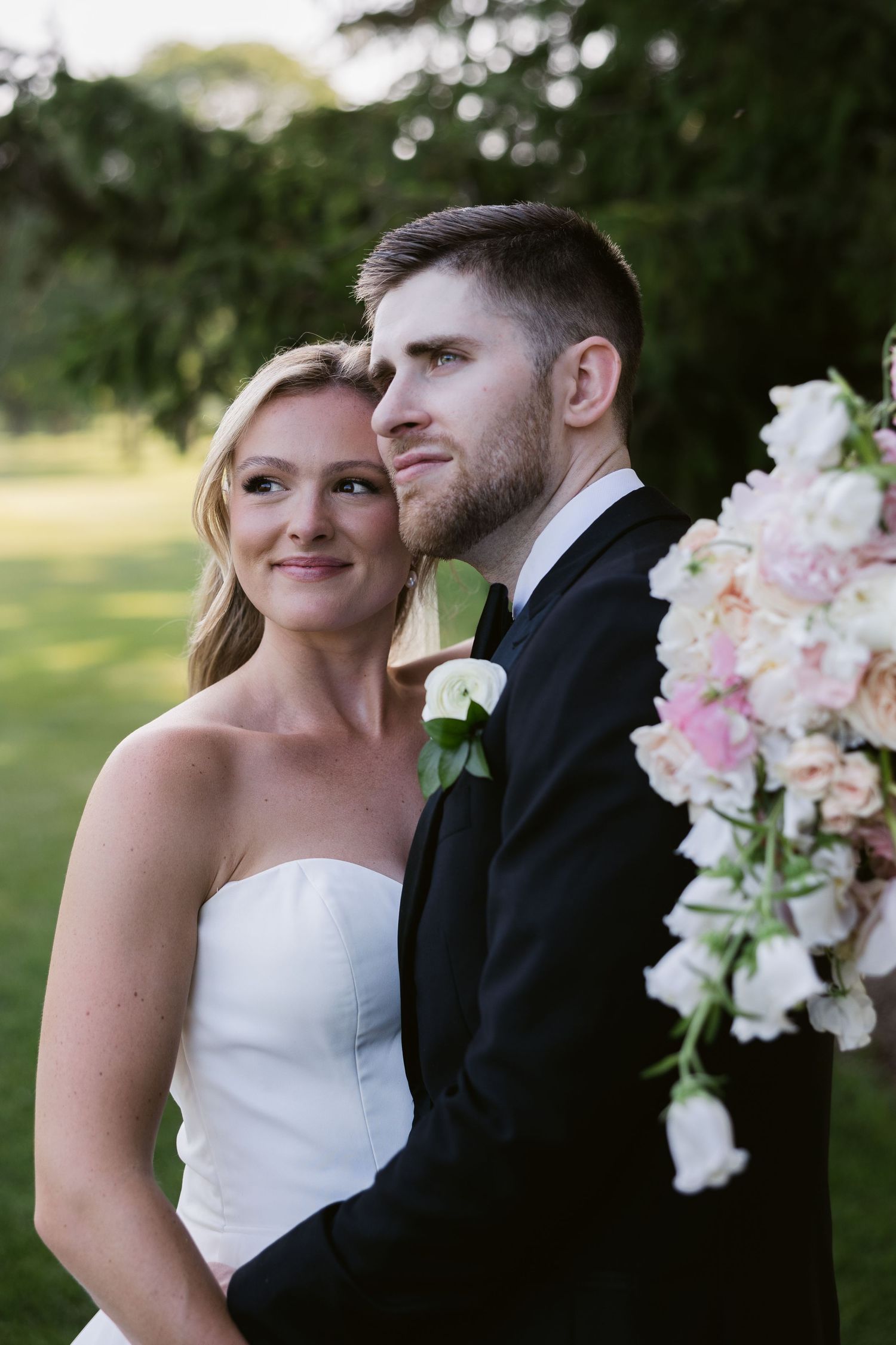 Wedding couple poses with pink and white floral bouquet against outdoor greenery.