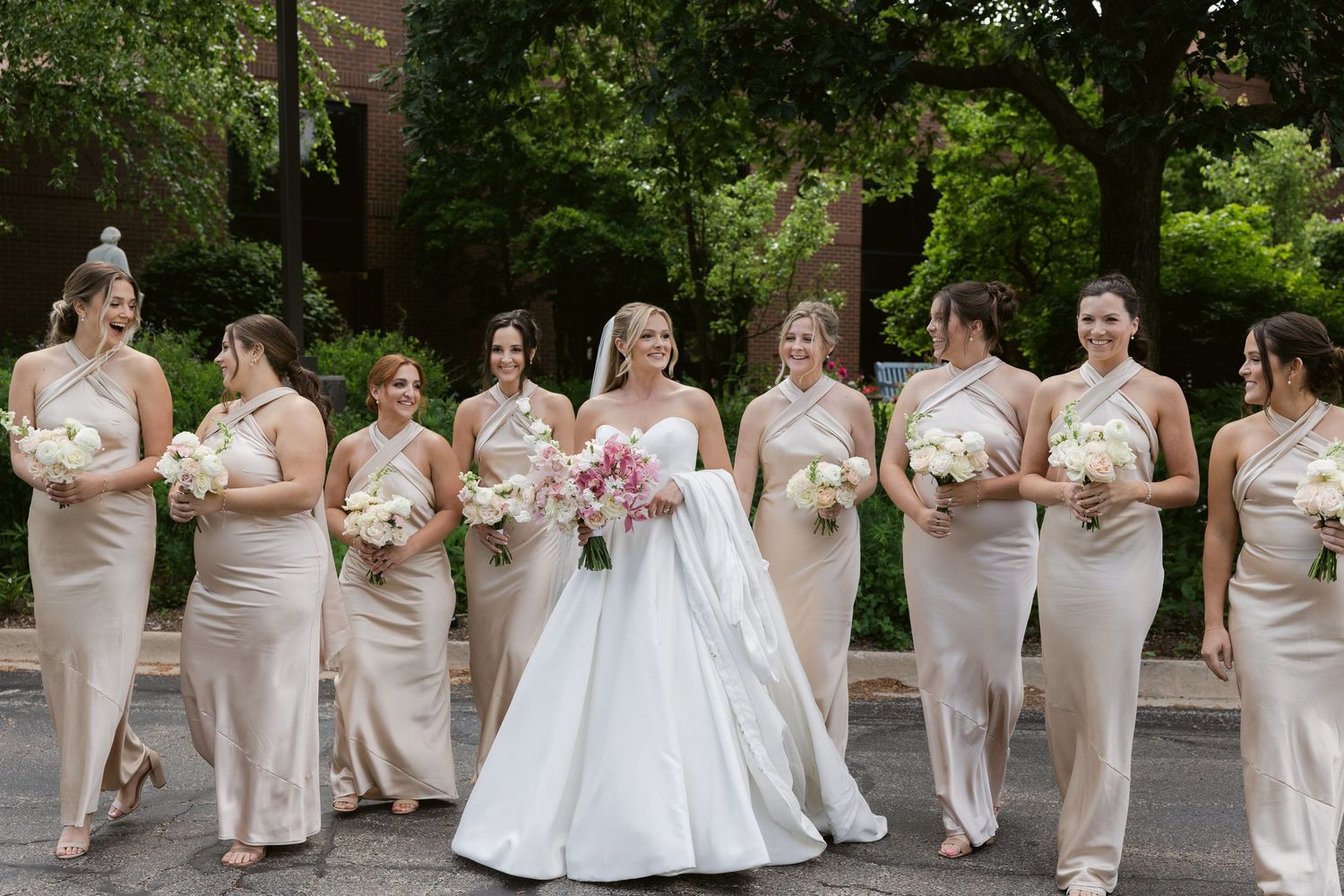 A wedding party in neutral dresses carries white and pink bouquets while walking outdoors.