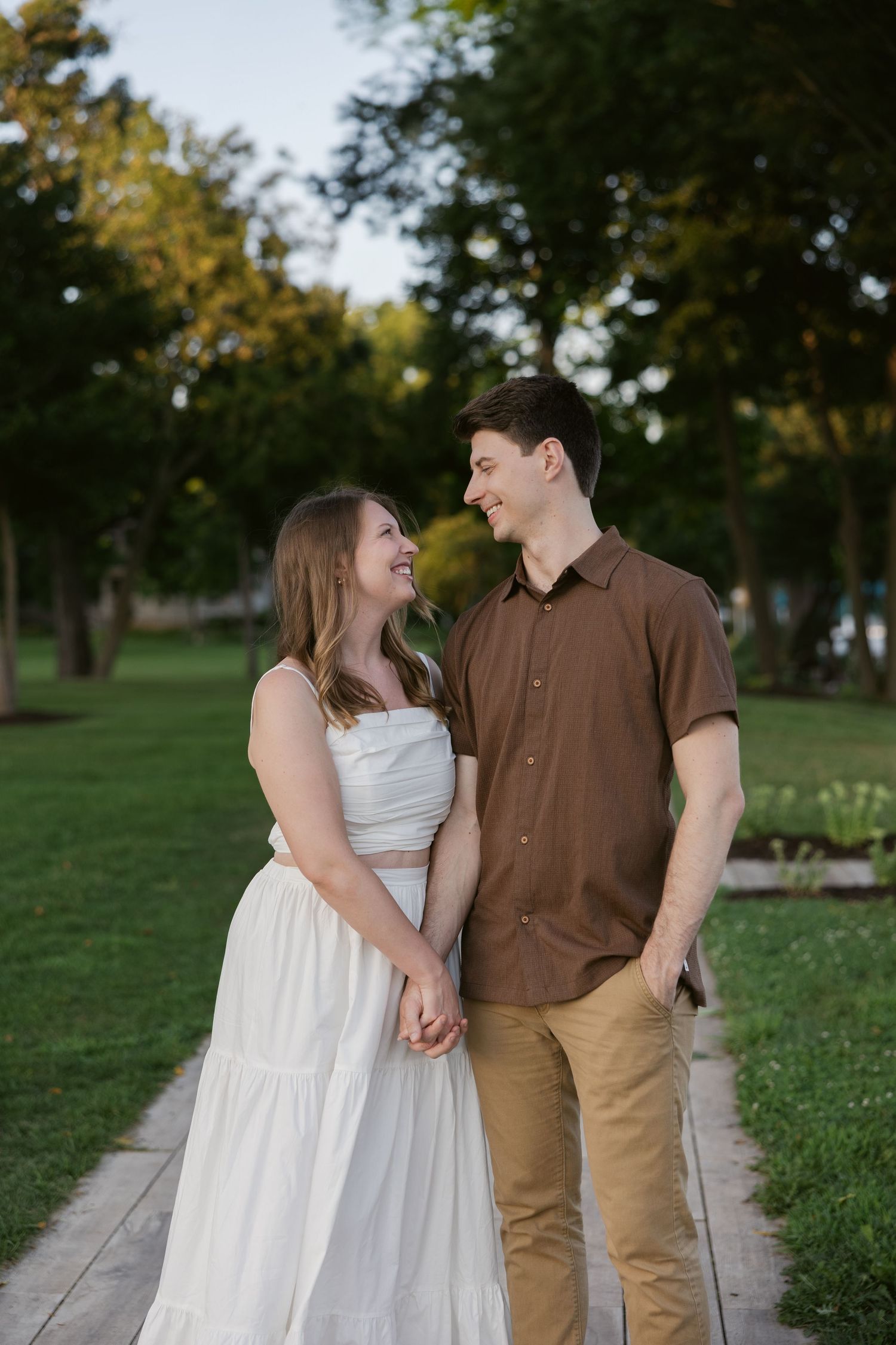 A couple holding hands and laughing together while standing on a wooden walkway in a park with green trees.