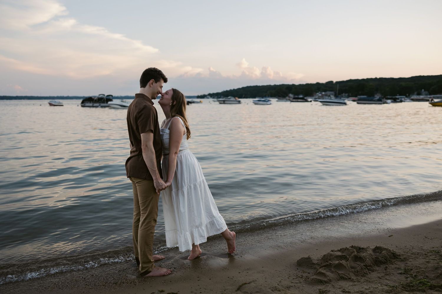 A couple enjoys a romantic moment along the shoreline at dusk with boats in the distance.