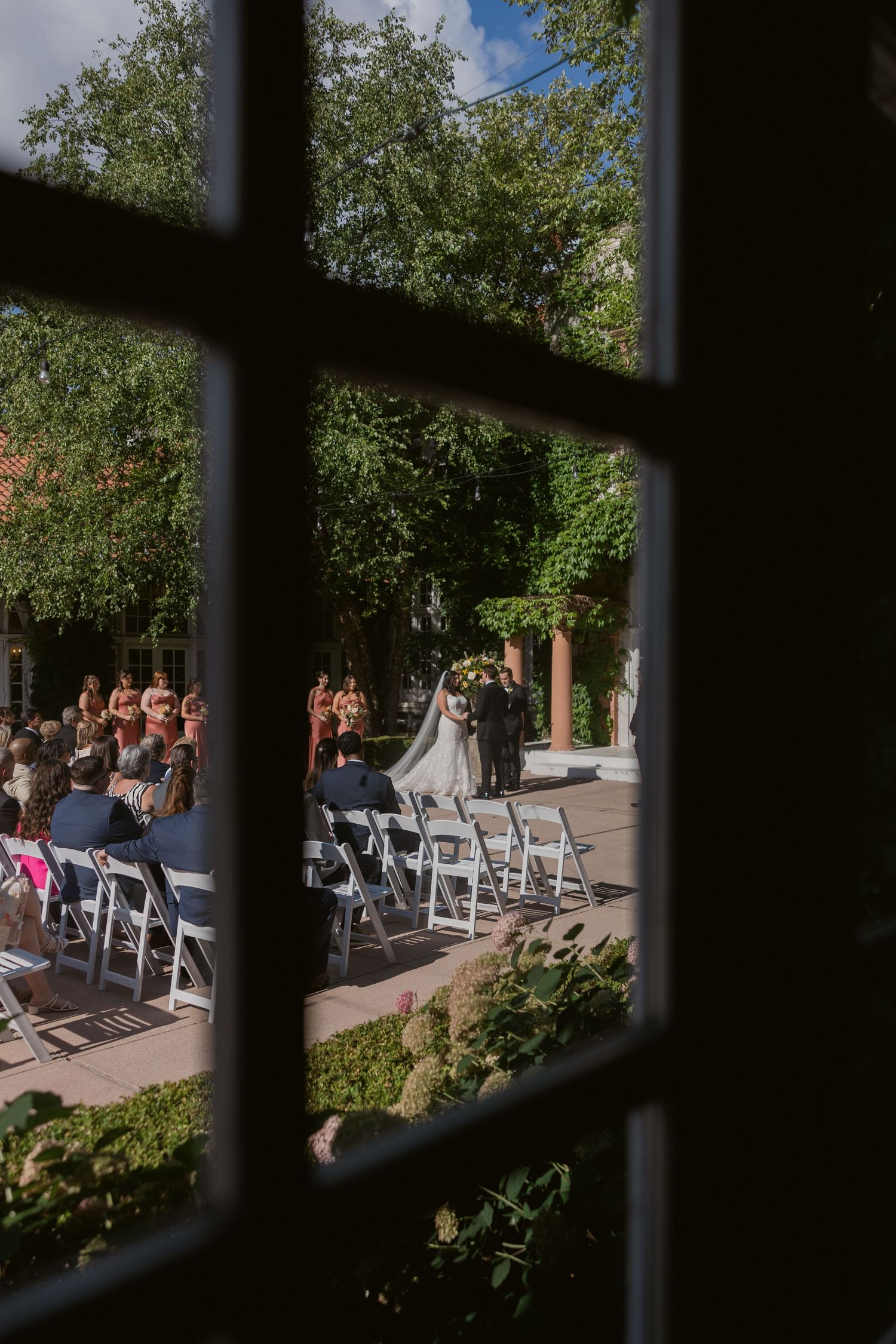 Wedding ceremony setup viewed through window panes with white chairs and guests on a sunny patio.