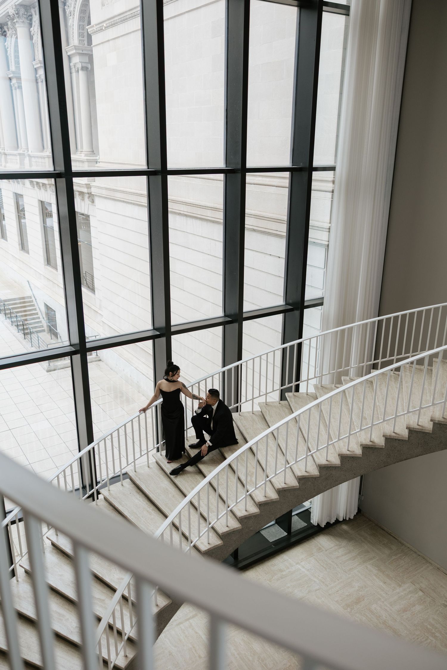 Couple sitting on white spiral staircase in modern architectural space with large windows.