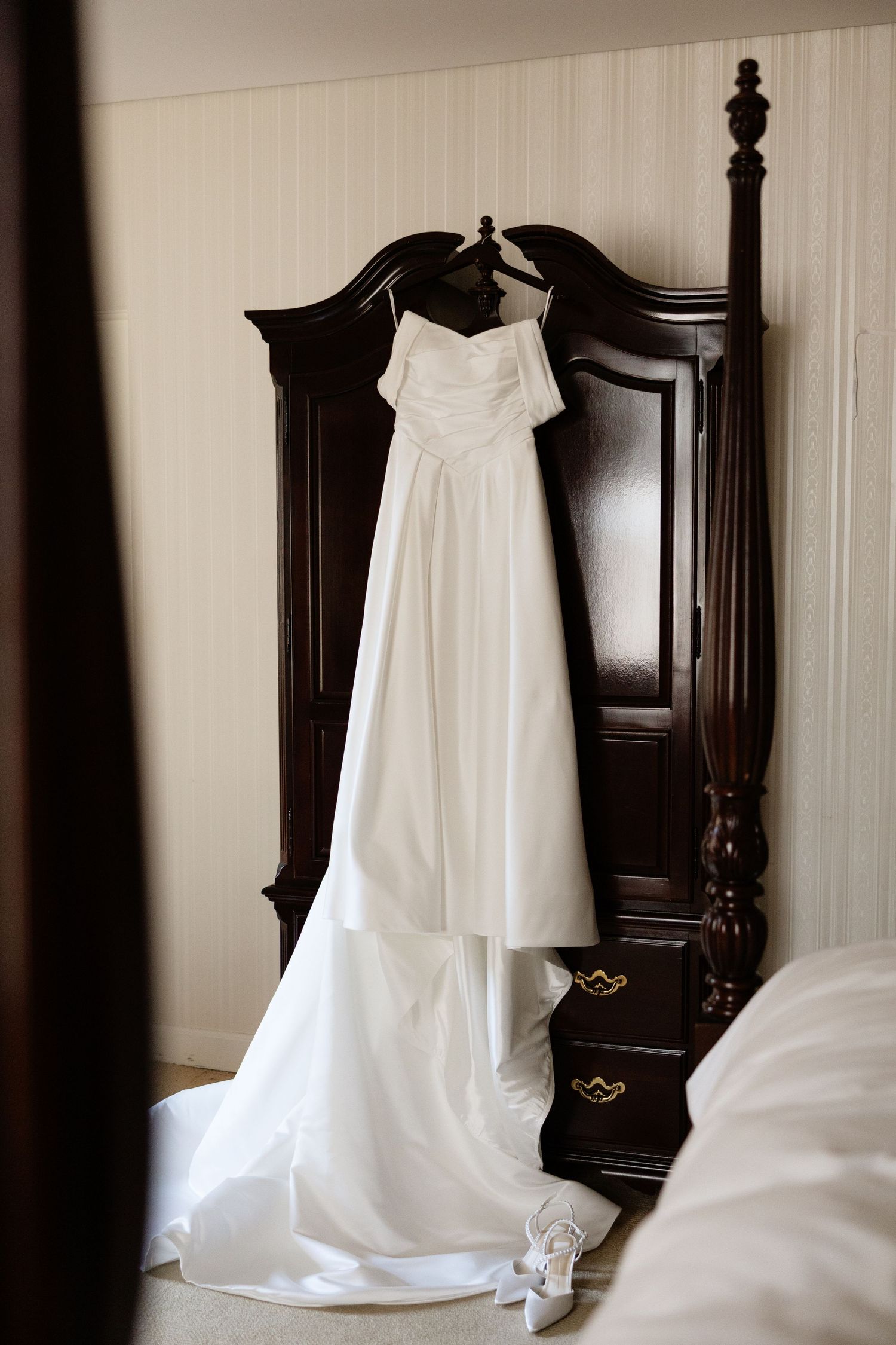 A white wedding dress hangs elegantly on a dark wooden bedpost, showcasing its flowing train and strapless design.