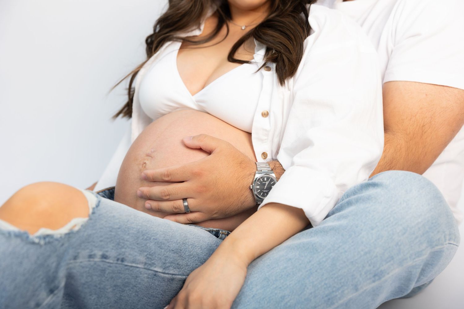Detail shot of hands cradling pregnant belly in casual denim and white outfit.