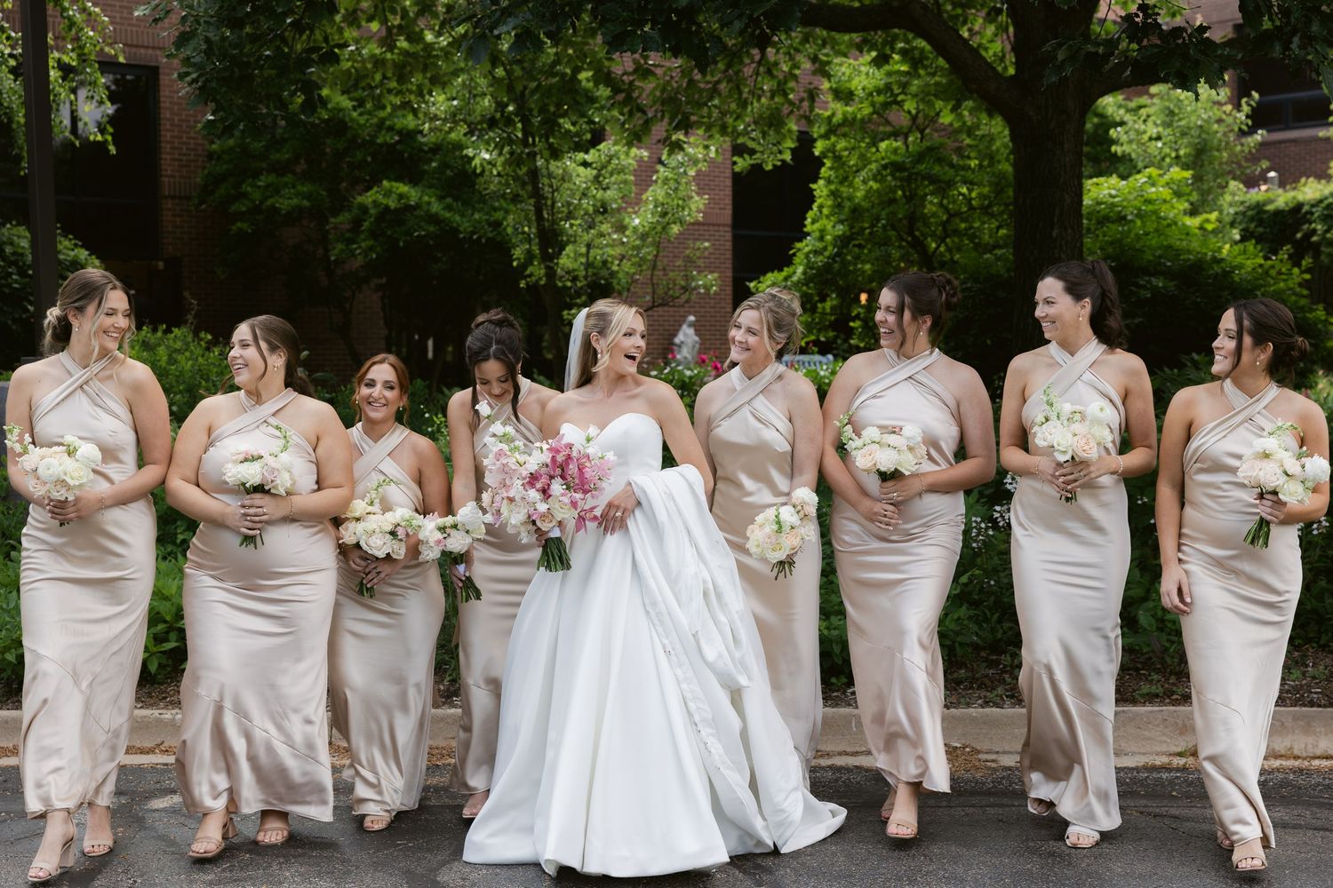 A panoramic wedding party group poses outdoors wearing white and champagne colored dresses with floral bouquets.