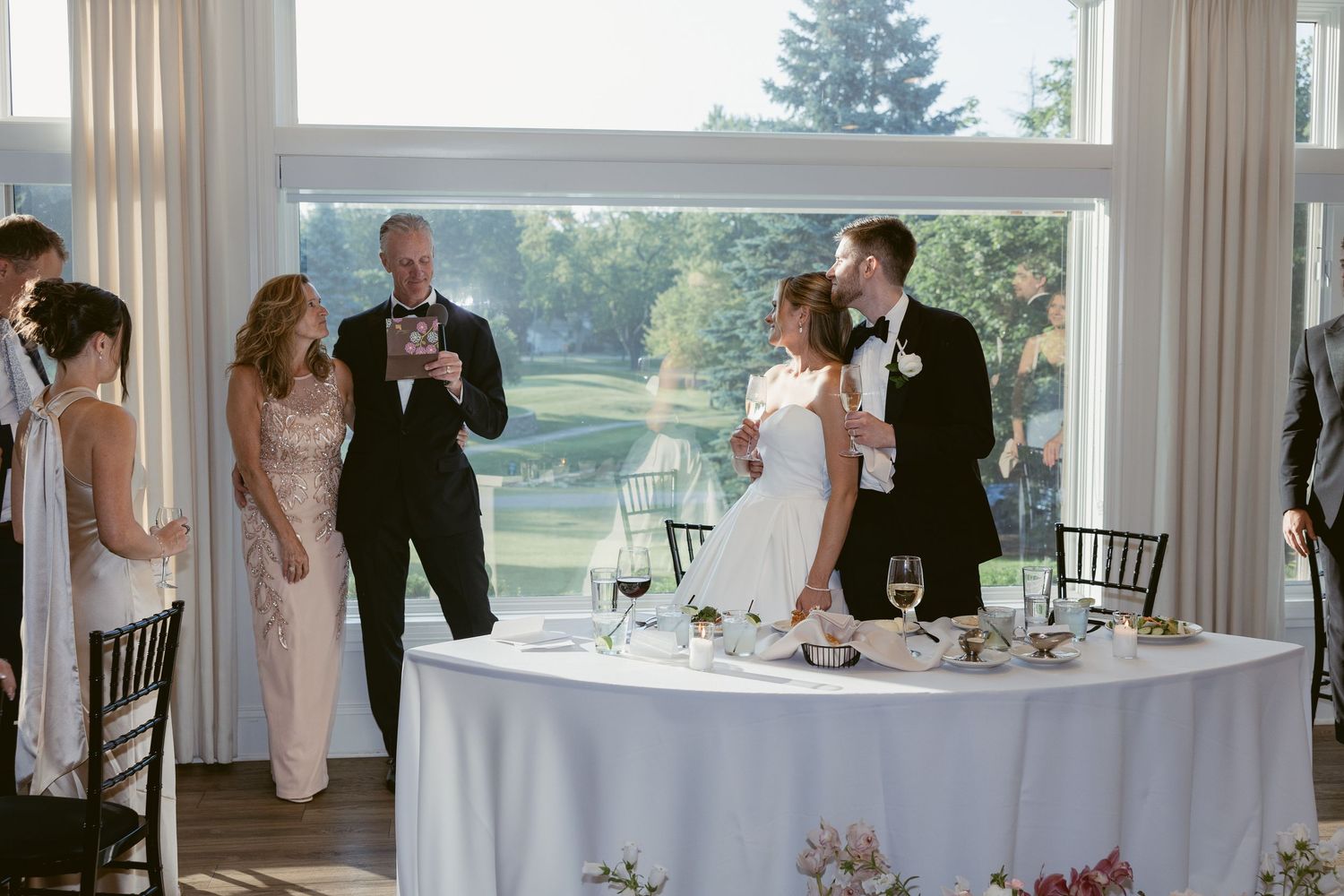Wedding reception scene with couple toasting at head table near large windows.