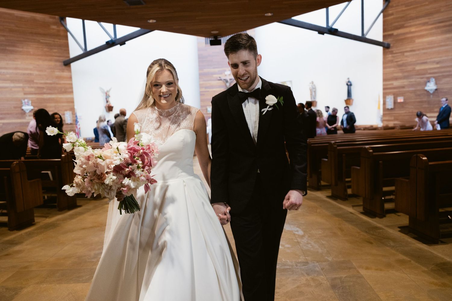 Newlyweds walking down wooden church aisle after ceremony with bride holding pink and white bouquet.