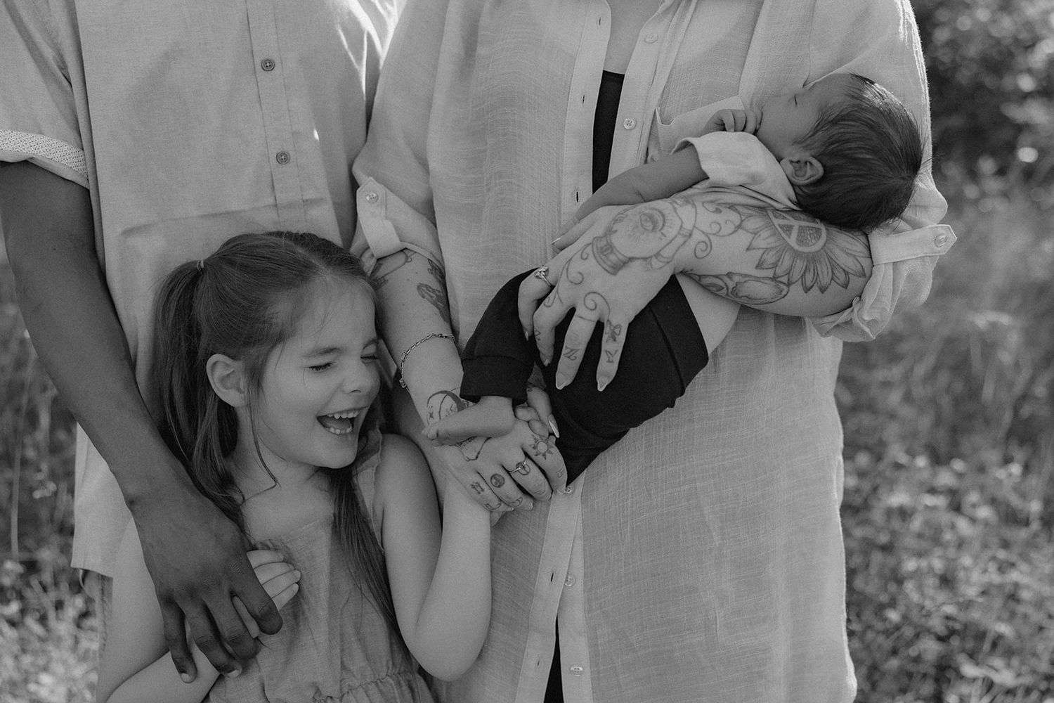 Family holds hands together in black and white photo with young child laughing outdoors.