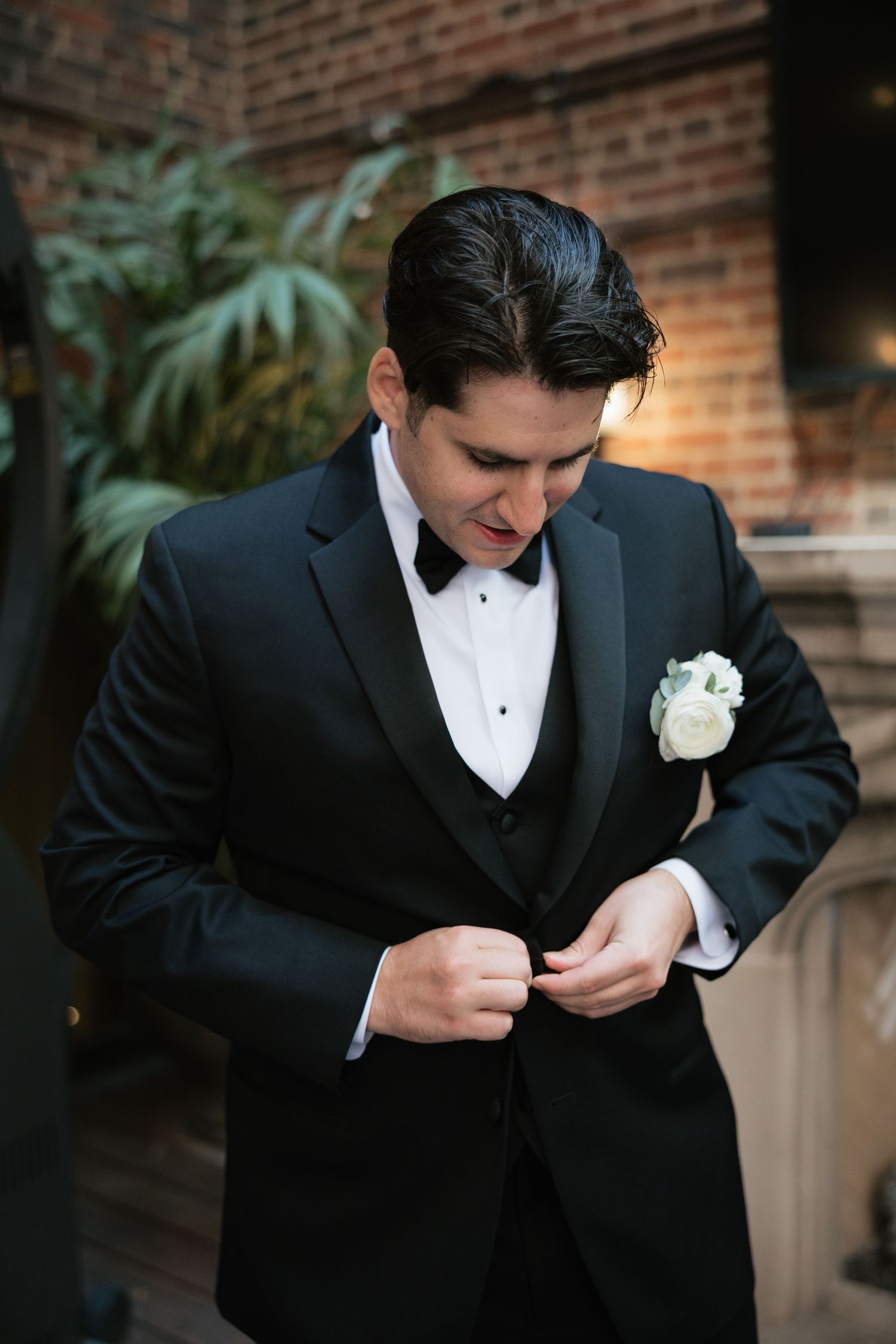 A groom in a black tuxedo adjusts his boutonniere against a brick wall background.