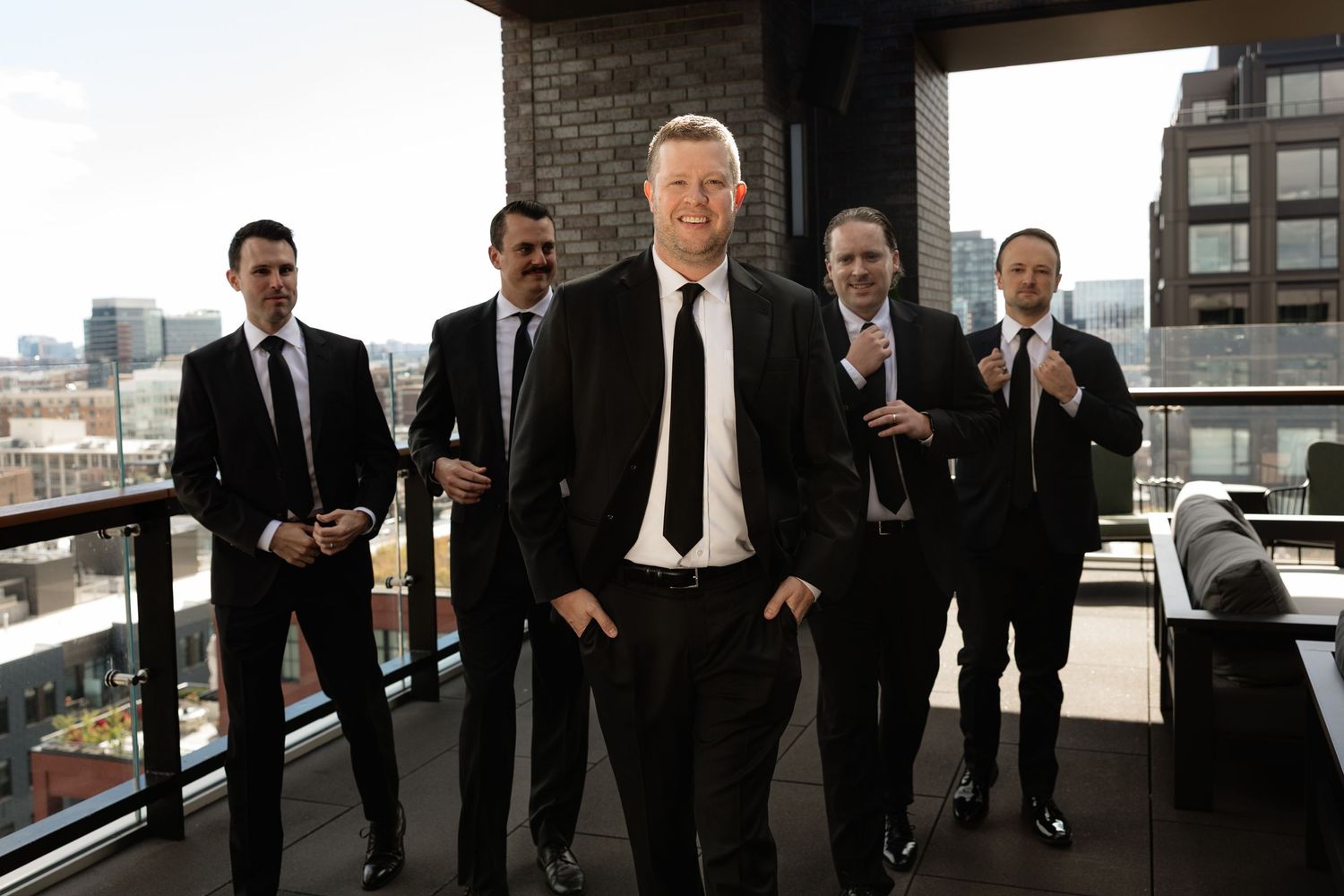 Groomsmen in black suits pose together in a modern urban setting with city views.