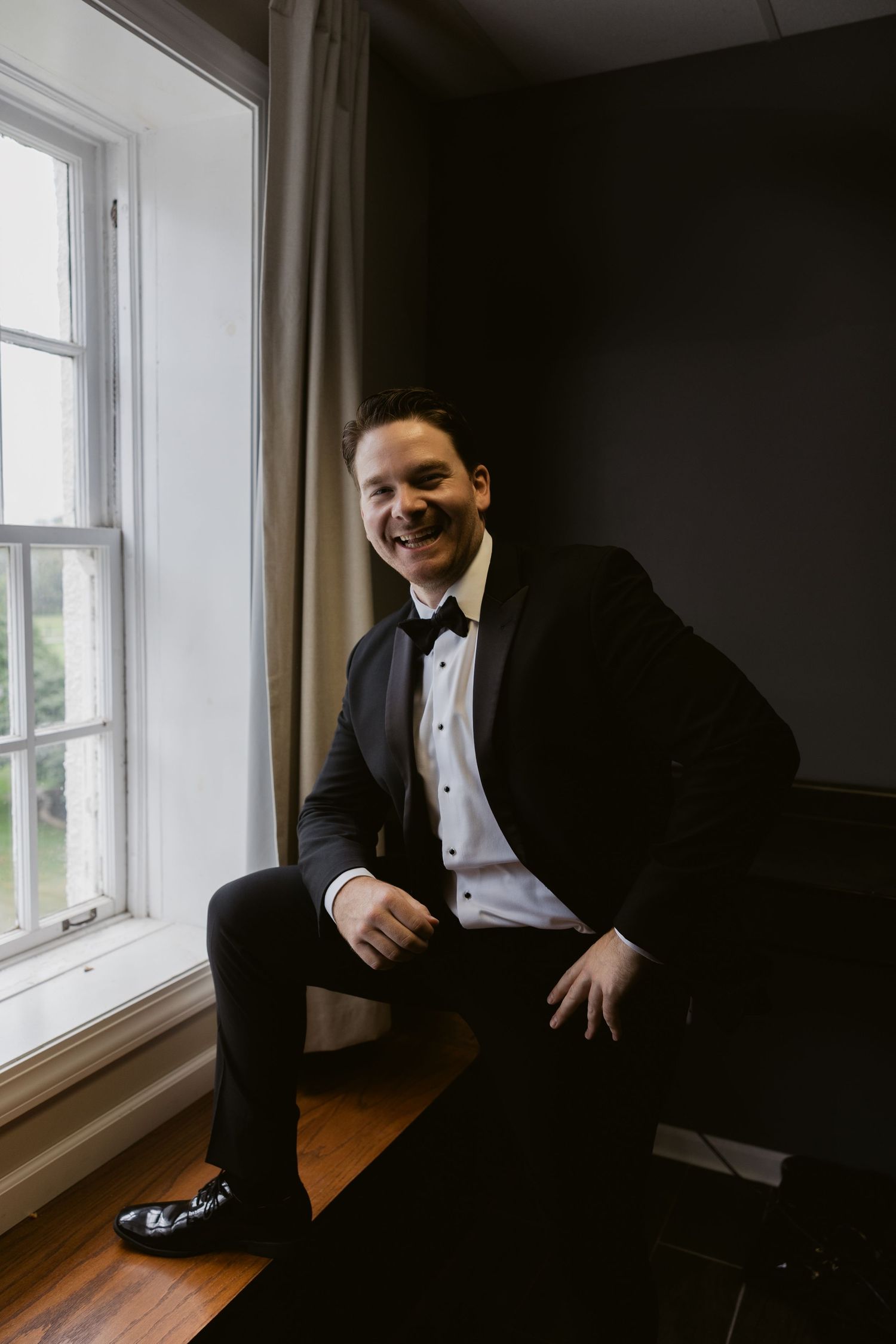A groom in a classic black tuxedo and bow tie sits casually by a window in a moody indoor setting.