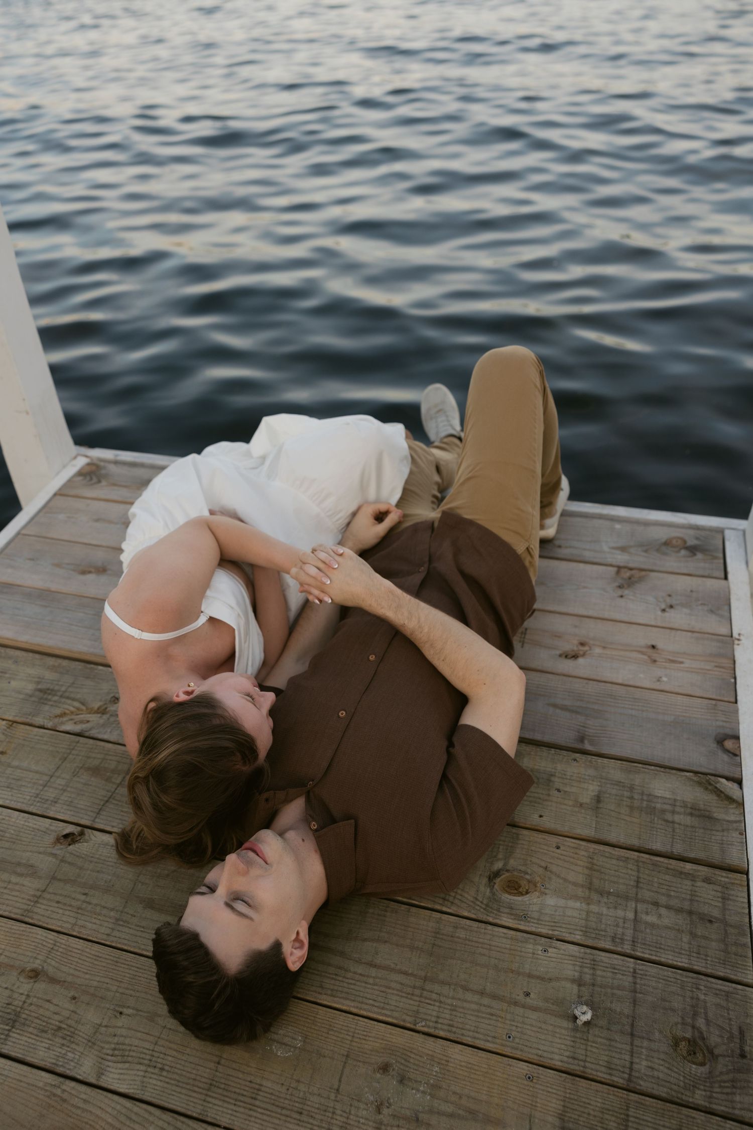 Romantic couple lying on wooden dock by water during sunset photoshoot.