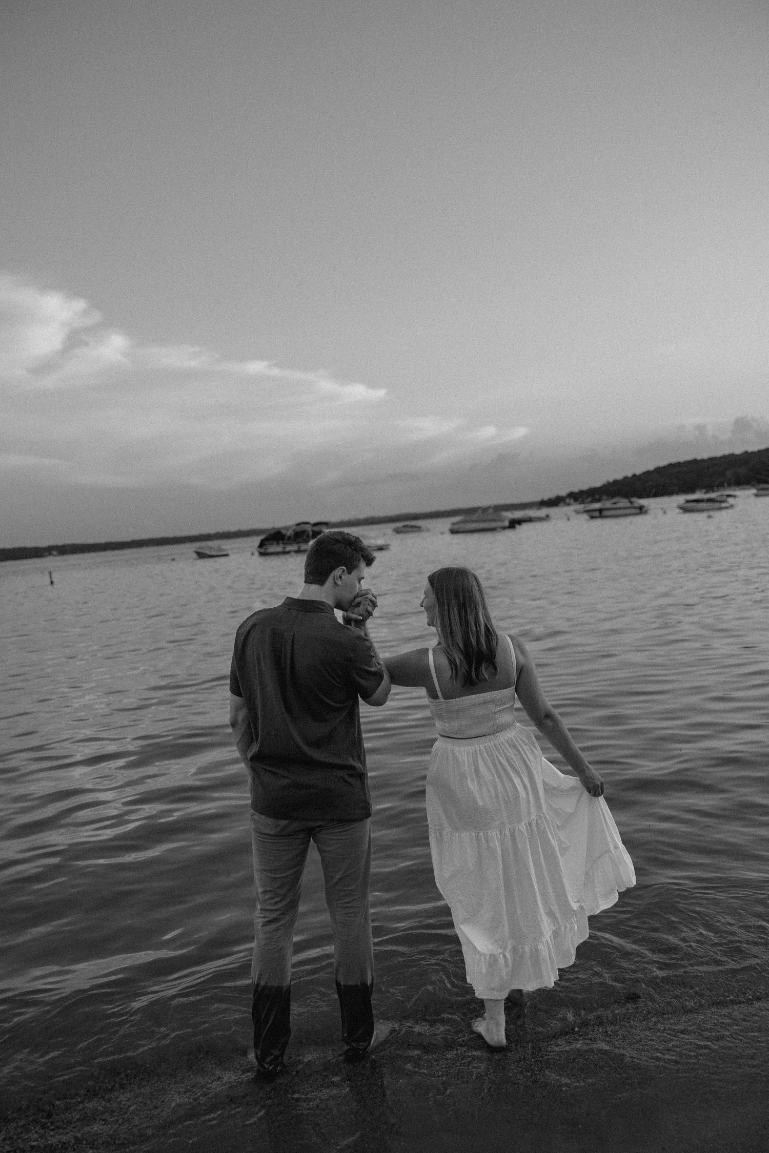 Two figures stand together by the ocean shore in dramatic black and white photography.