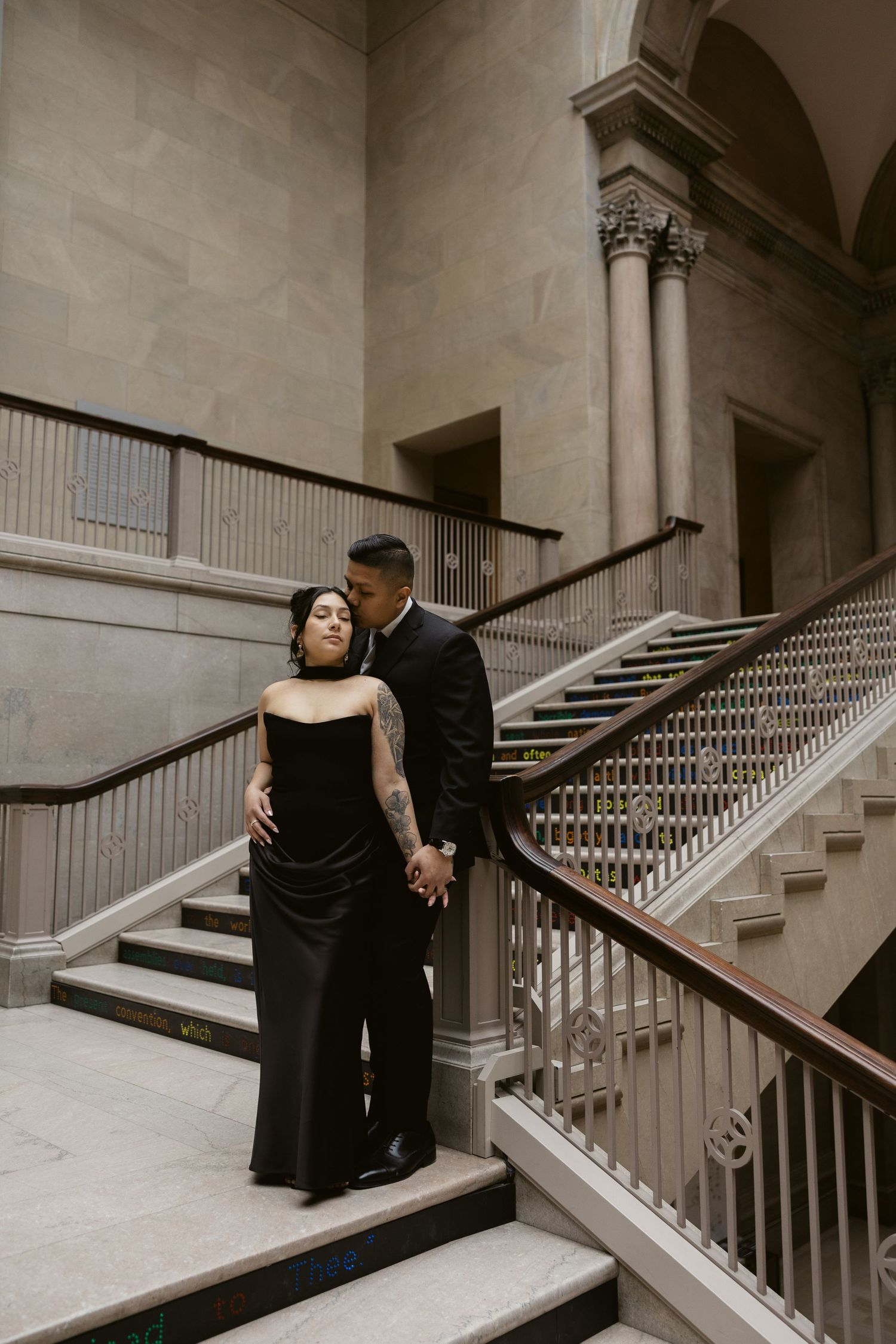 A couple in formal black attire embraces on an elegant marble staircase in a historic building.