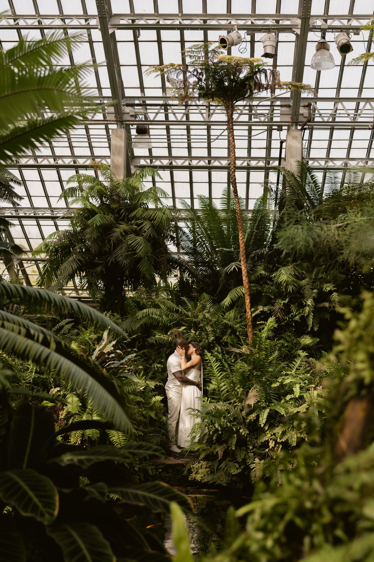 White-clad figures share an intimate moment among verdant greenhouse foliage and glass paneled ceilings.