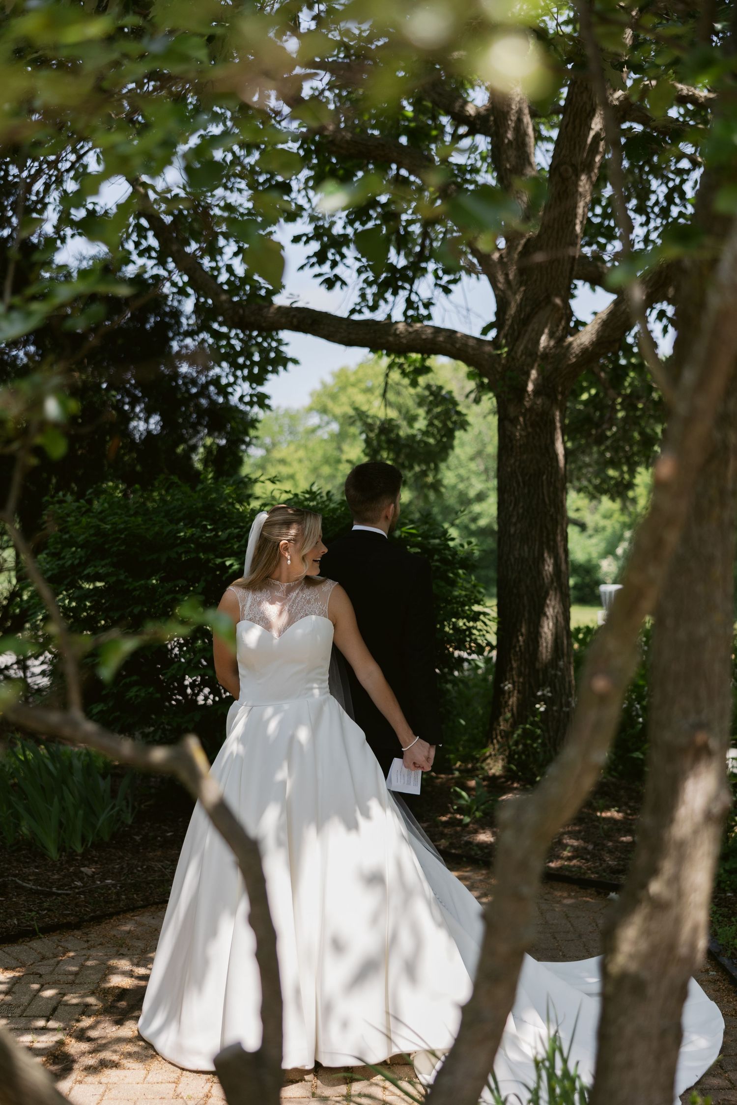 Wedding couple walking among trees with dappled sunlight creating beautiful shadows on white dress.